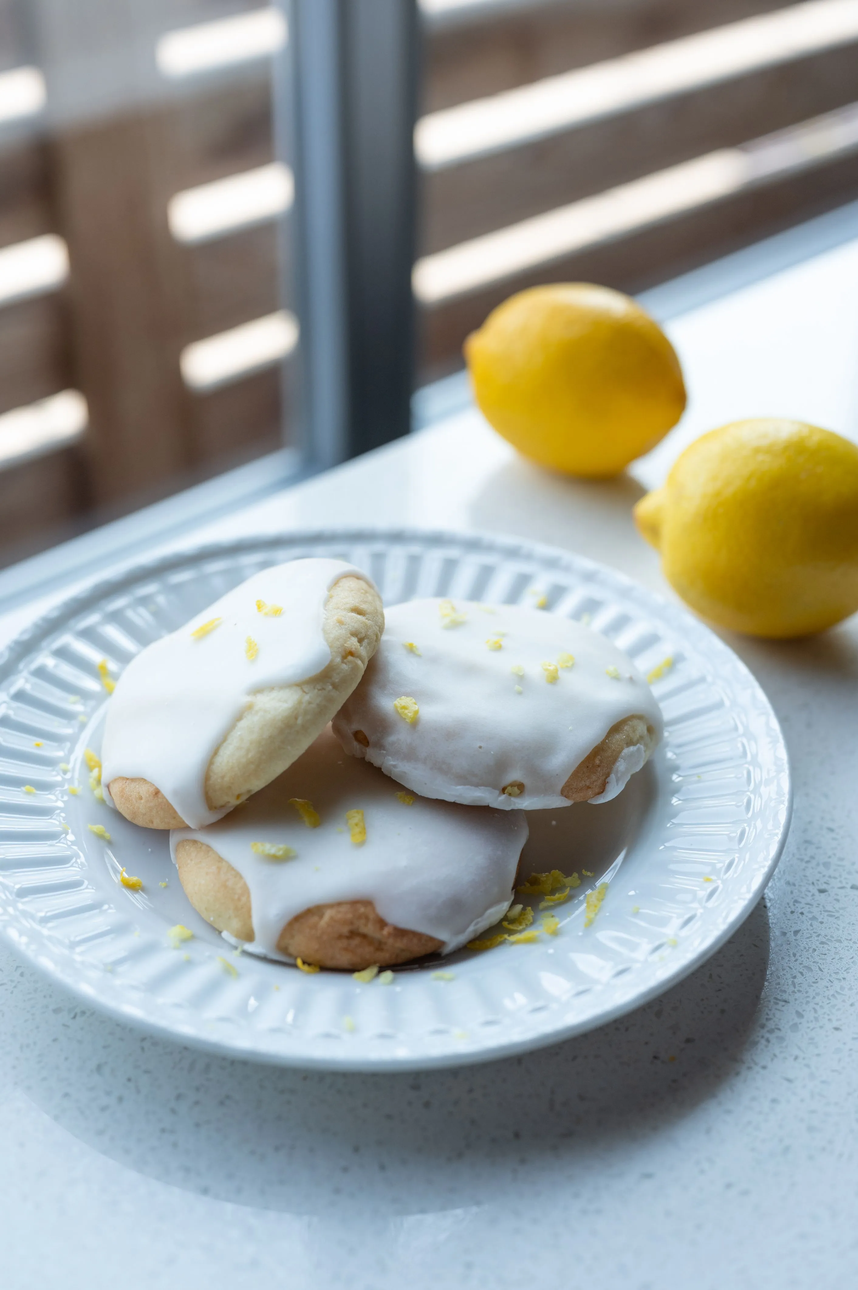 Lemon cookies with white icing and zest on a decorative plate, with fresh lemons in the background.