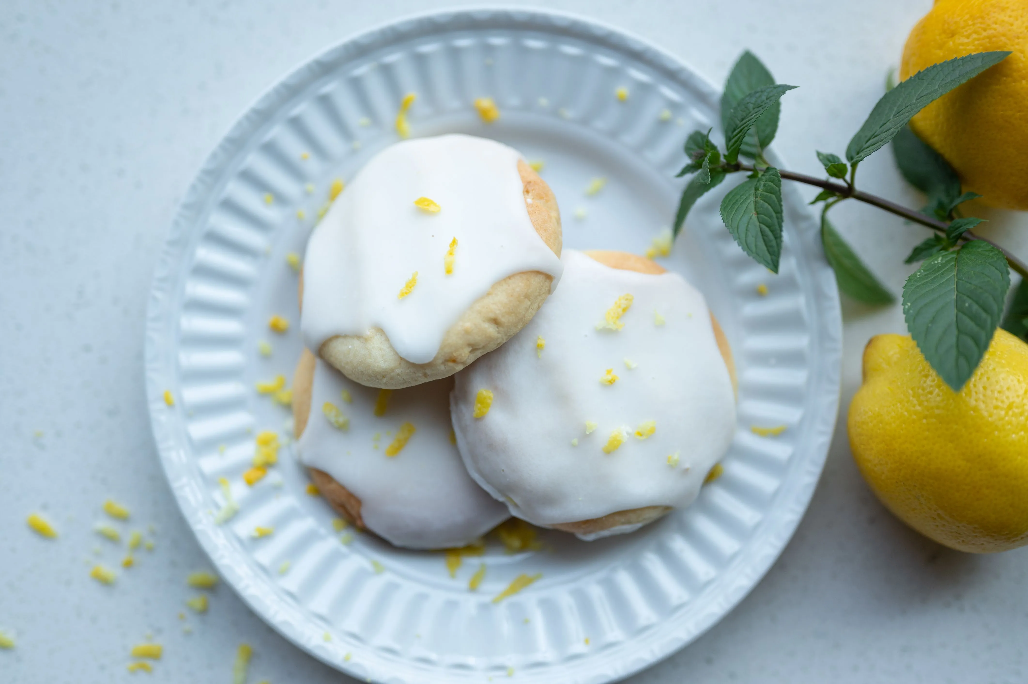 Plate of lemon cookies with white icing and lemon zest on top, next to fresh lemons and mint leaves.
