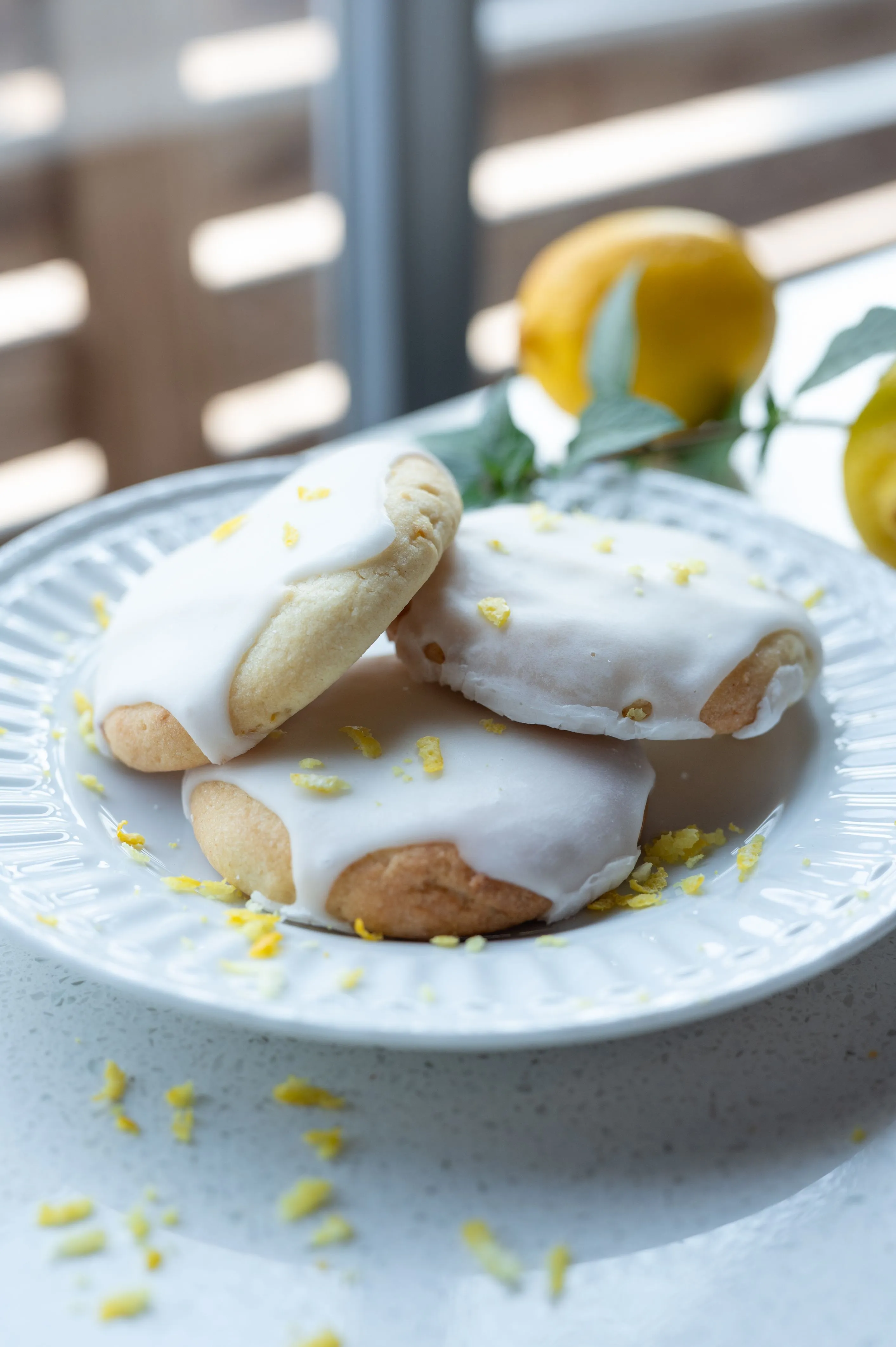 Lemon cookies with white icing on a plate, garnished with lemon zest, with lemons and leaves in the background.