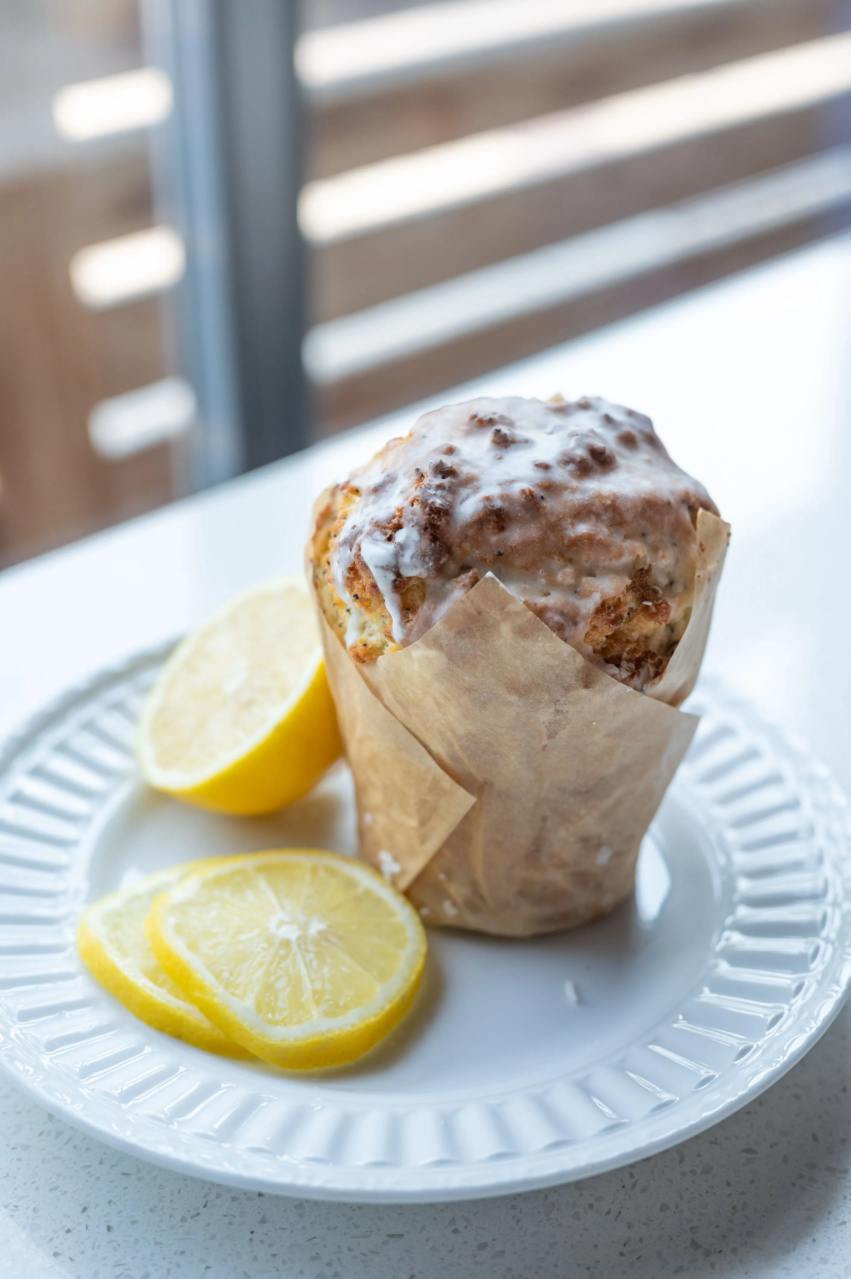 Lemon poppy seed muffin with a glazed top on a white plate with lemon slices, near a sunlit window.