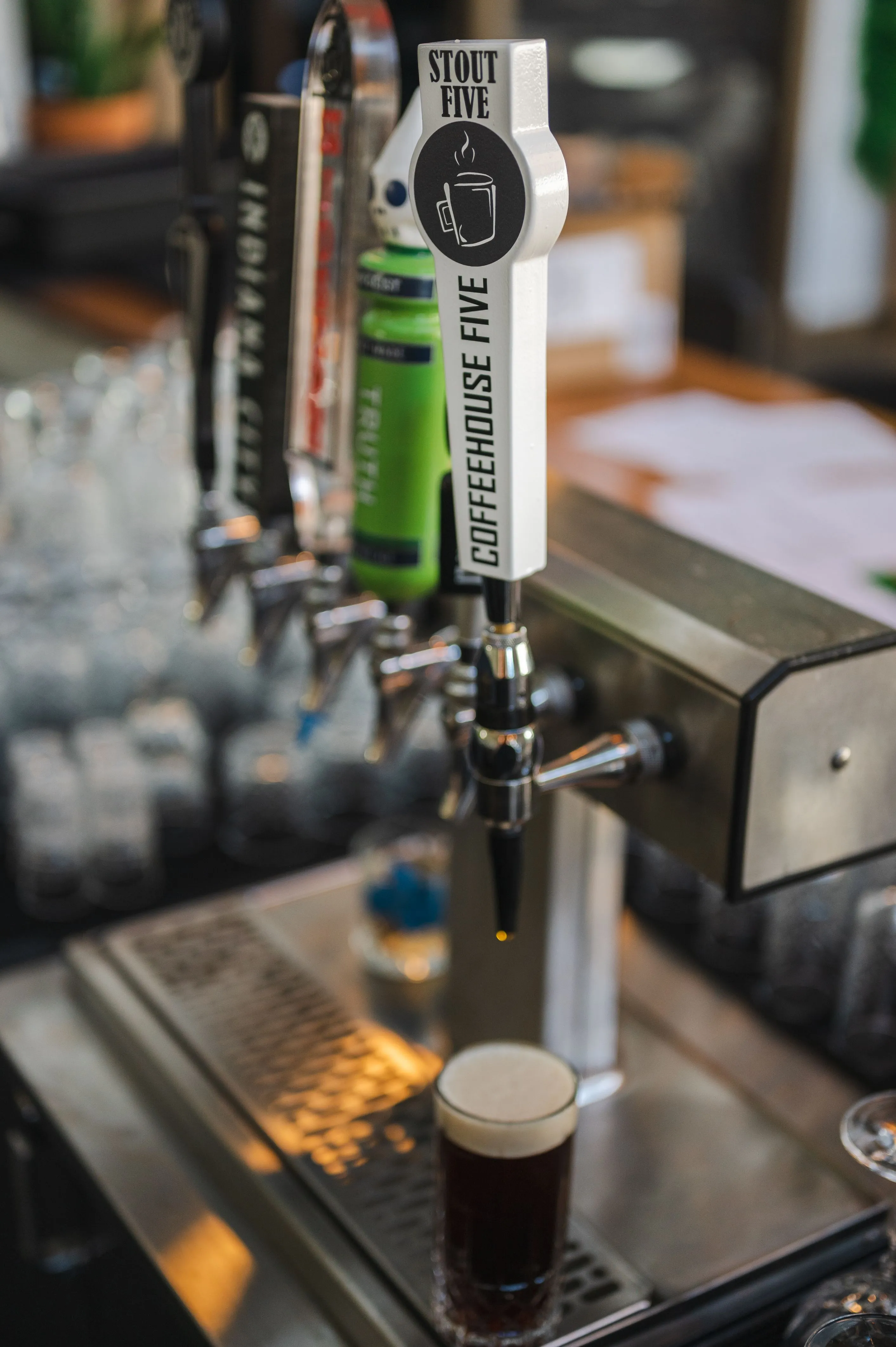 Close-up of a row of beer taps at a bar, with one featuring a "STOUT FIVE COFFEEHOUSE" label, and a pint of dark beer with a frothy head standing on the counter.