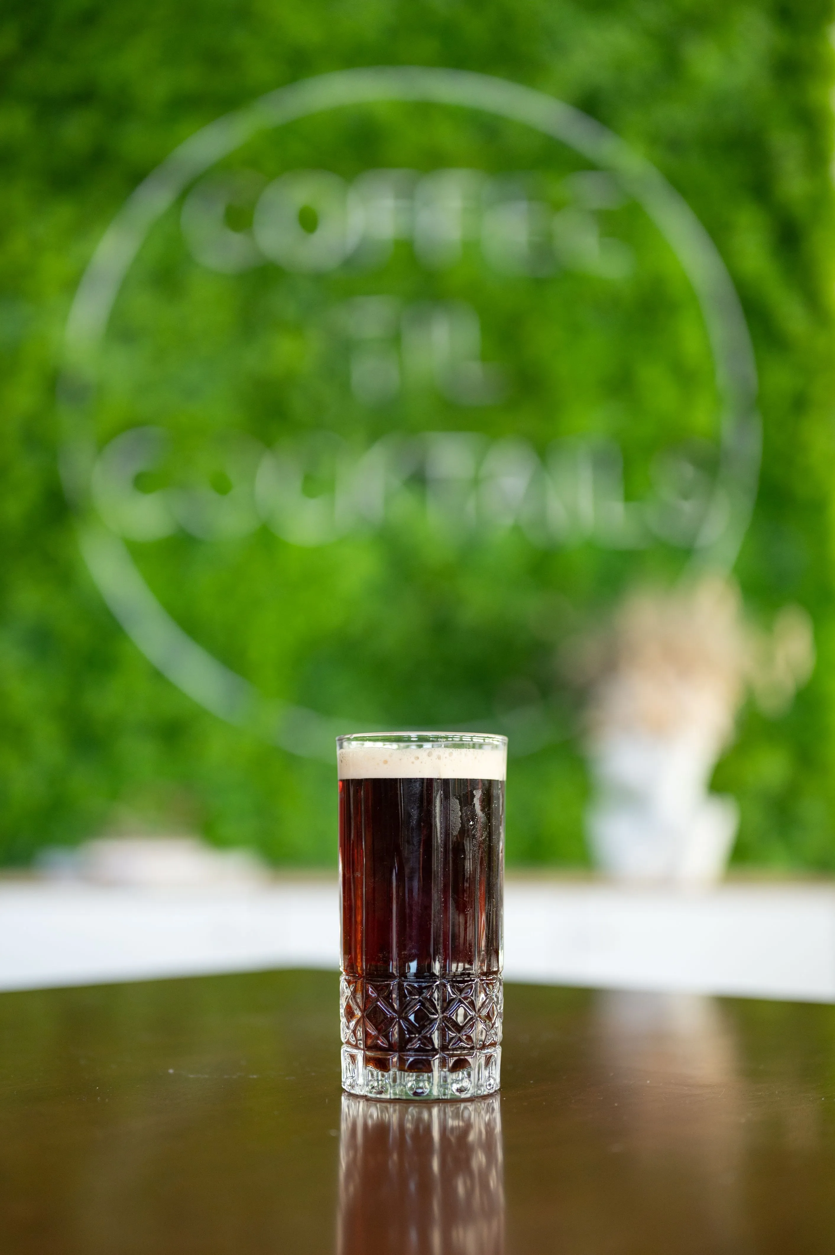 A glass of dark beer on a shiny table with an out-of-focus green background displaying the words "Coffee till Cocktails".