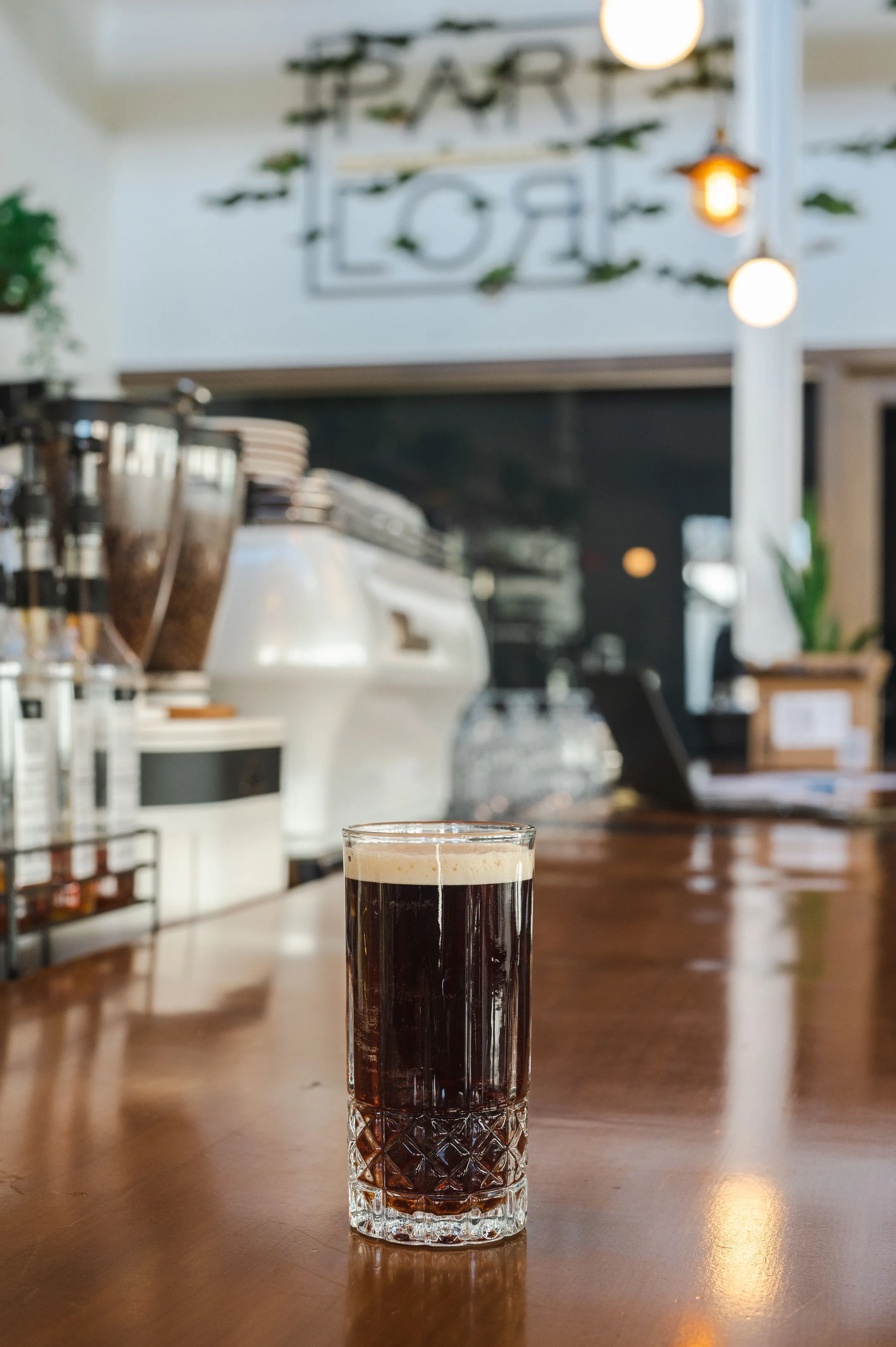 A cold brew coffee with a creamy foam on top, on a wooden counter in a cafe with blurred espresso machines and decor in the background.