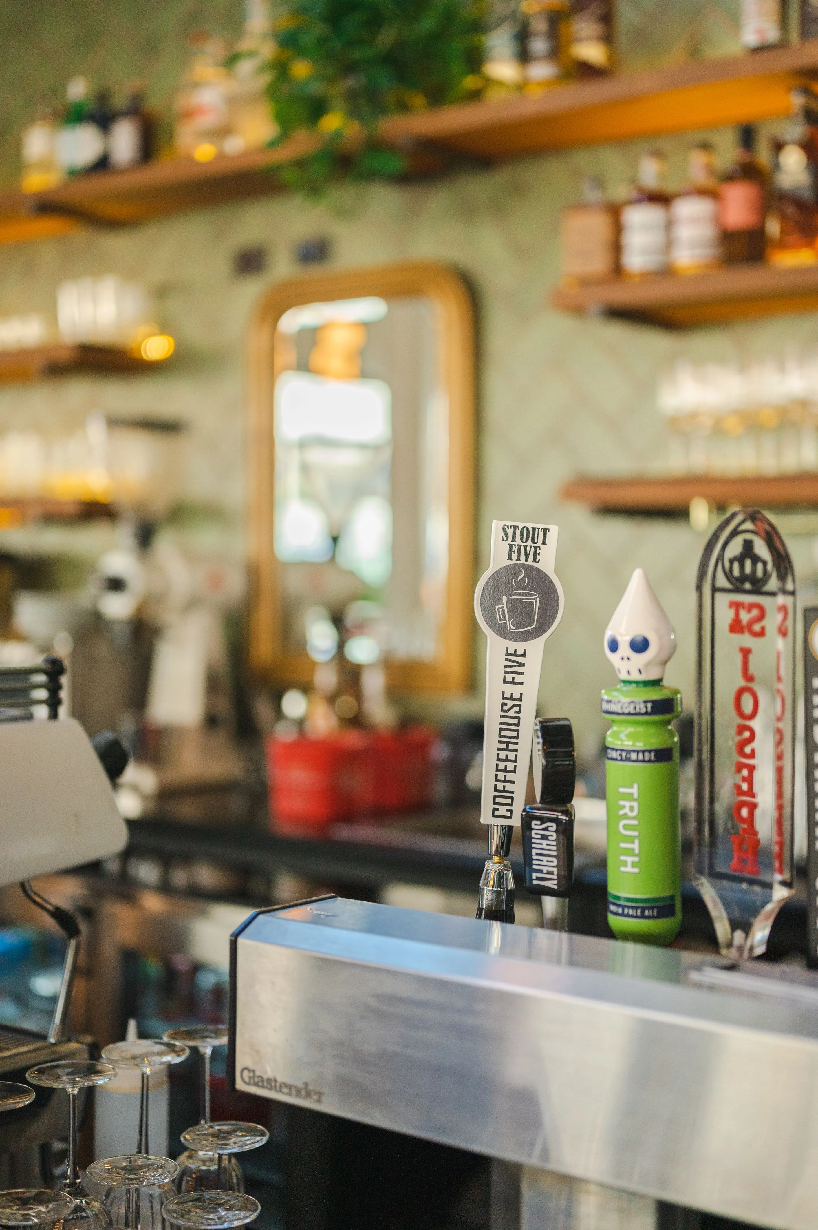 A cozy cafe bar featuring a series of tap handles, including a stylized stout dispenser, with a blurred background highlighting cafe shelves, a mirror, and warm lighting.