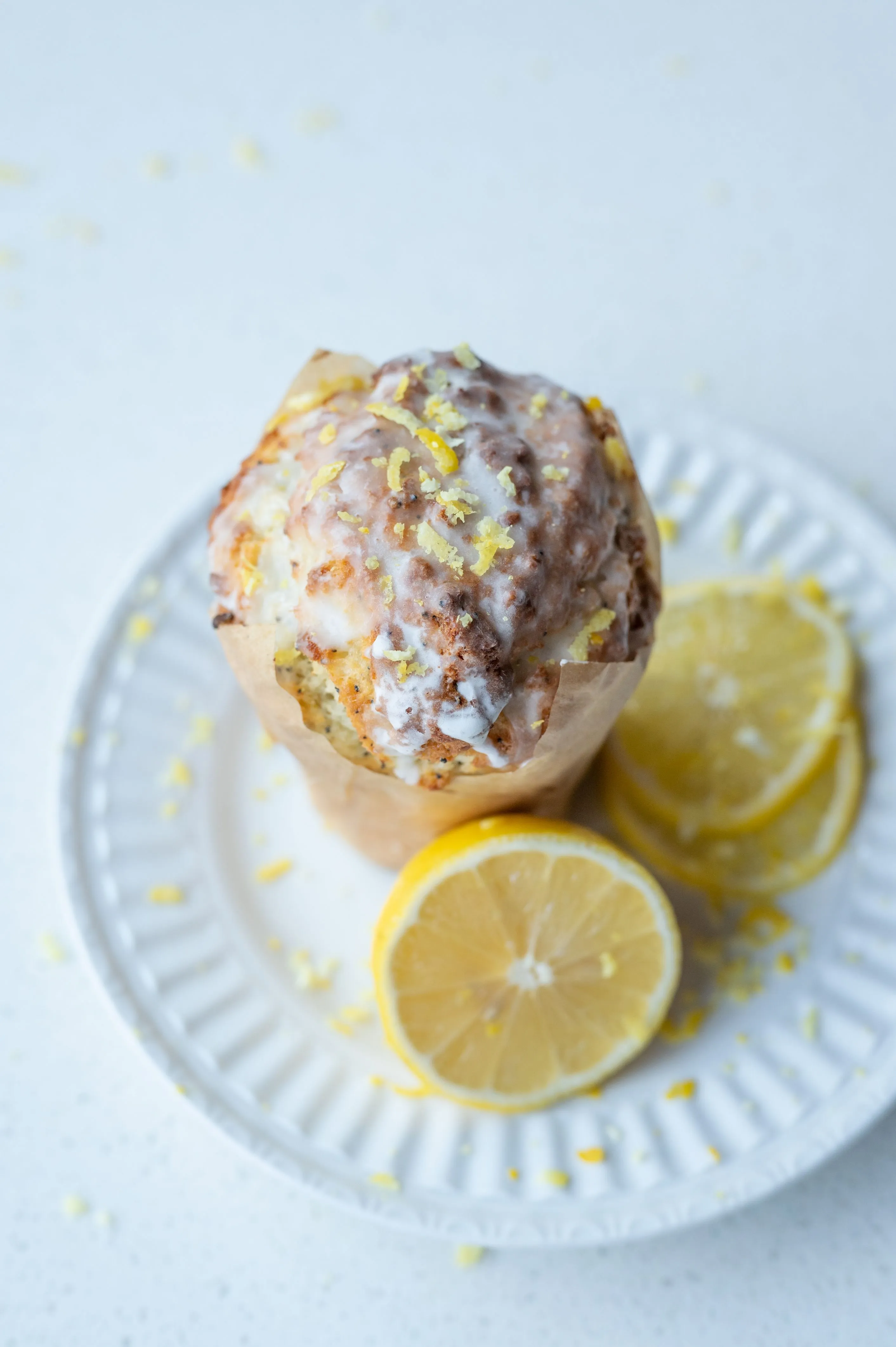 Lemon poppy seed muffin with glaze and lemon zest on a white plate with lemon slices and zest around, on a light surface.
