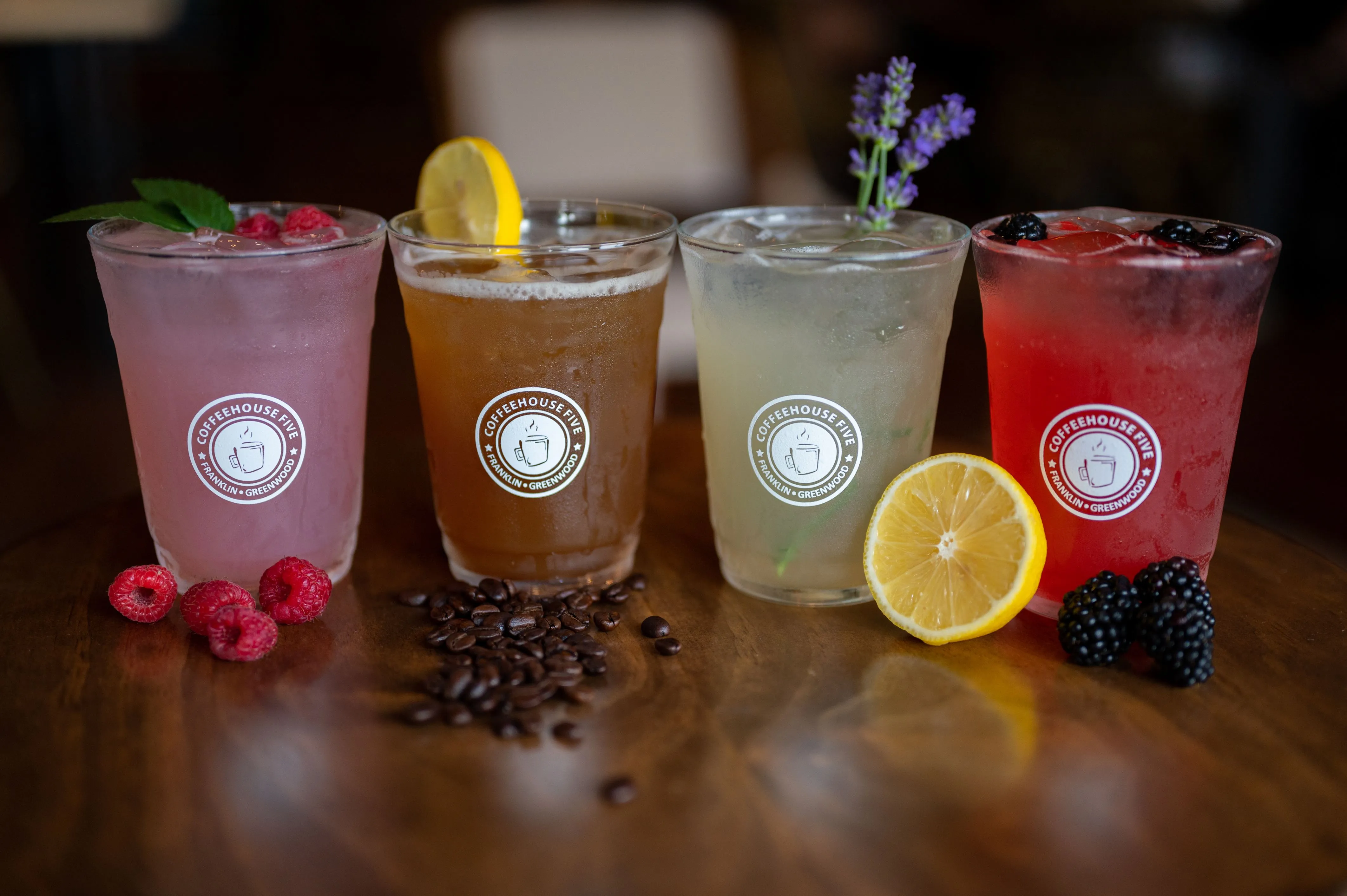 Four specialty drinks in branded glasses, garnished with fresh fruits and coffee beans on a wooden table.