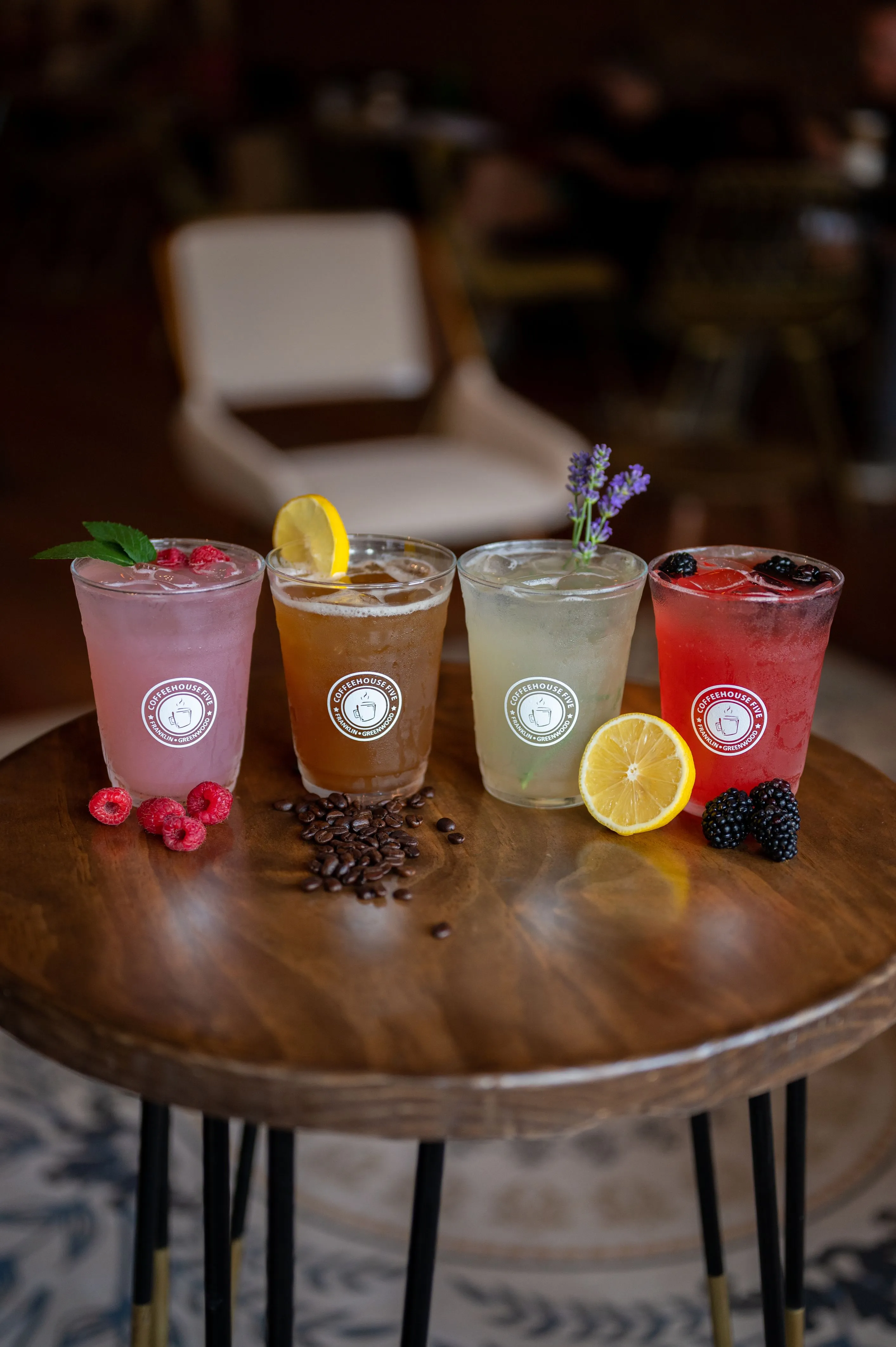 Four assorted flavored iced beverages on a wooden table, each garnished with different items such as raspberries, a lemon slice, lavender, and blackberries, with scattered coffee beans on the table.