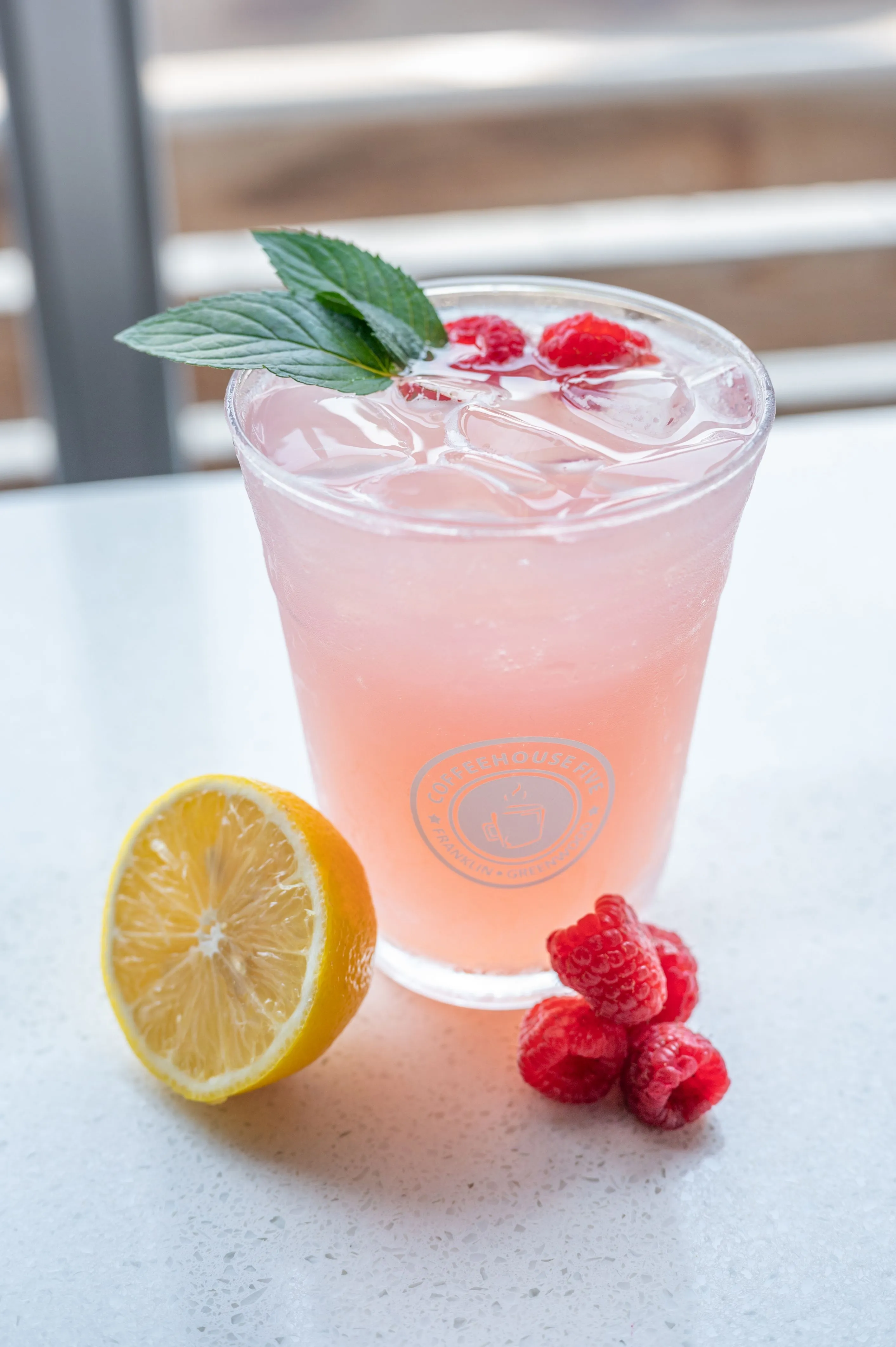 Refreshing pink lemonade in a glass with ice and raspberries, garnished with a mint leaf, accompanied by half a lemon and whole raspberries on a marble surface.