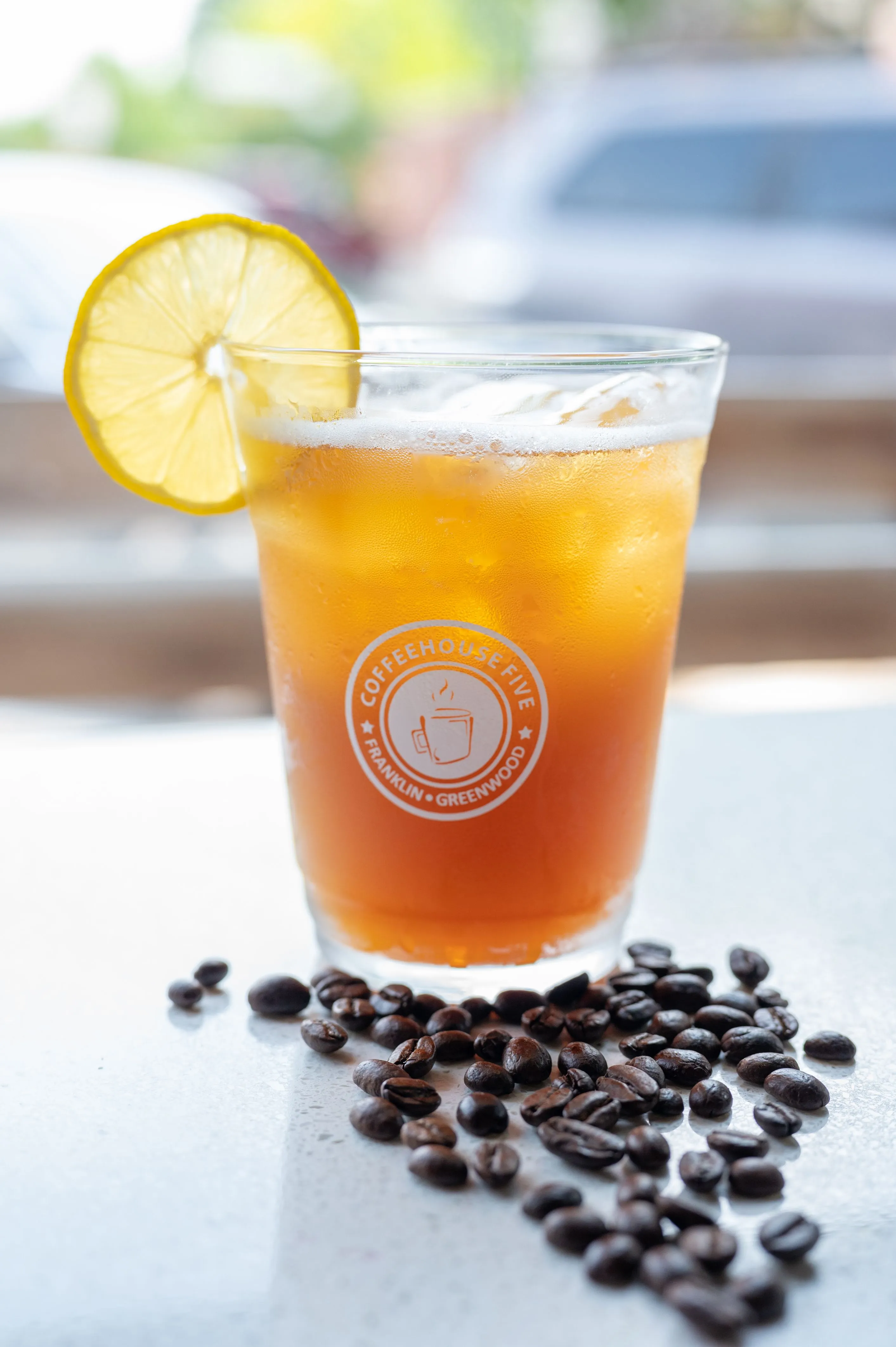 Iced coffee in a glass with a logo, garnished with a lemon slice, surrounded by coffee beans on a table, with a blurred background.
