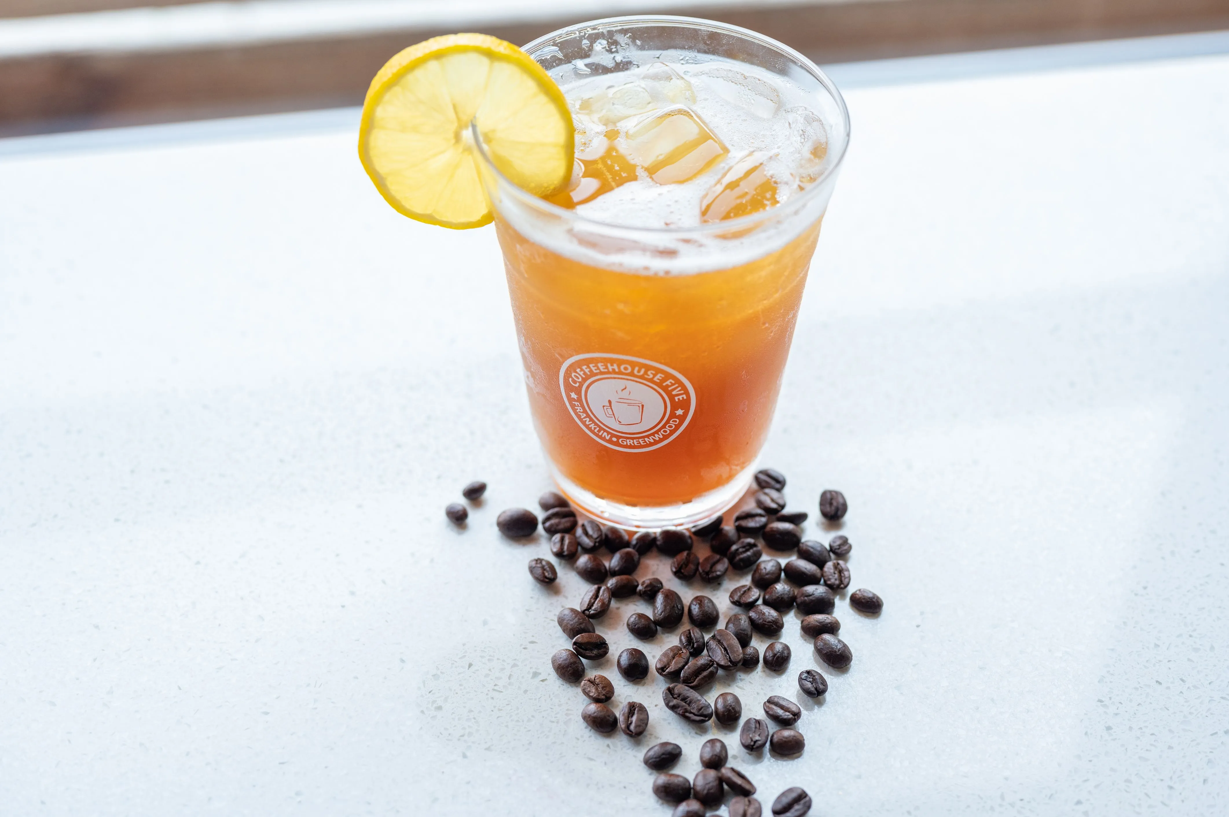 Iced coffee beverage garnished with a lemon slice, surrounded by scattered coffee beans on a light surface.
