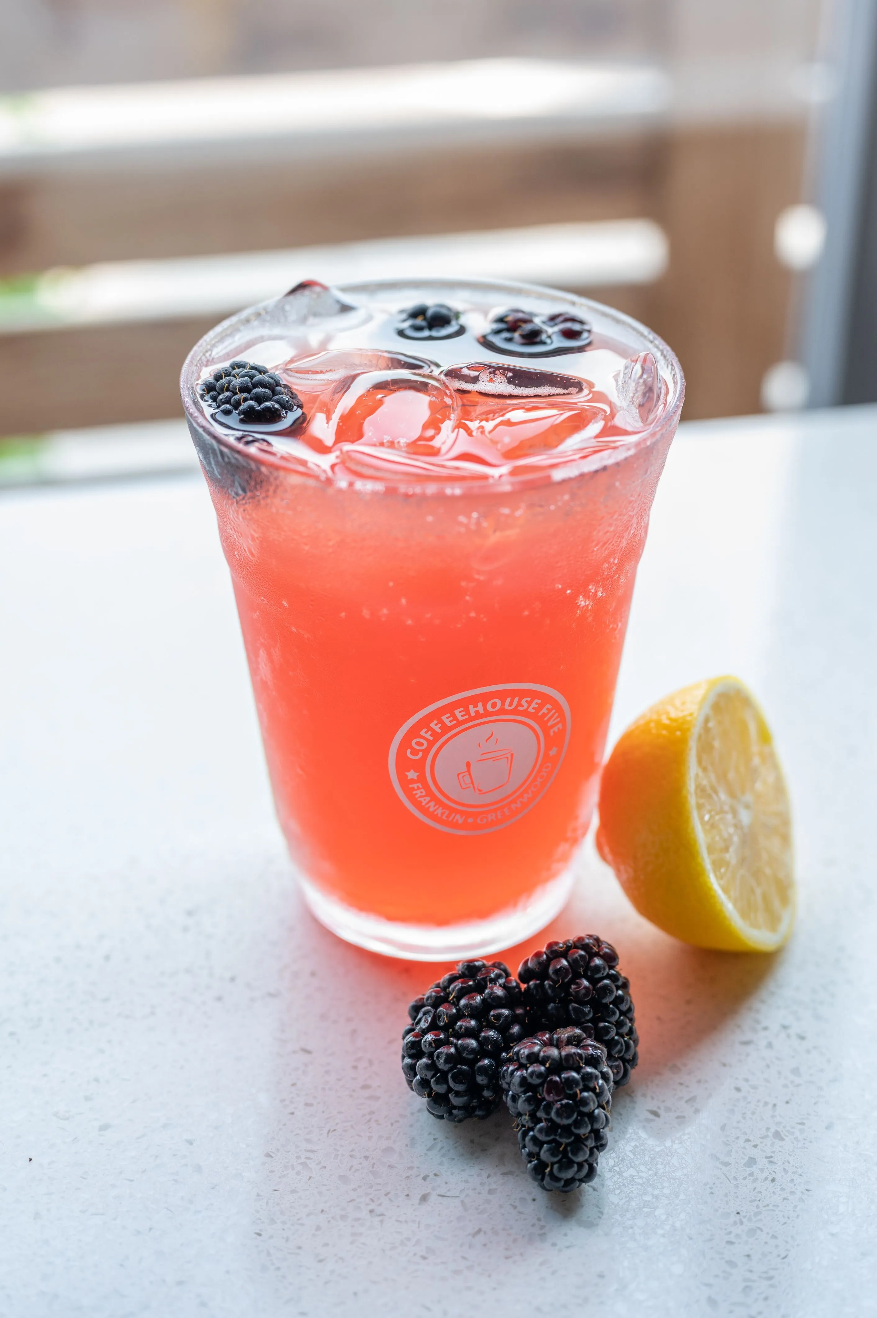 Iced pink beverage garnished with blackberries, served with a lemon slice and extra blackberries on the side, with visible logo on the glass.