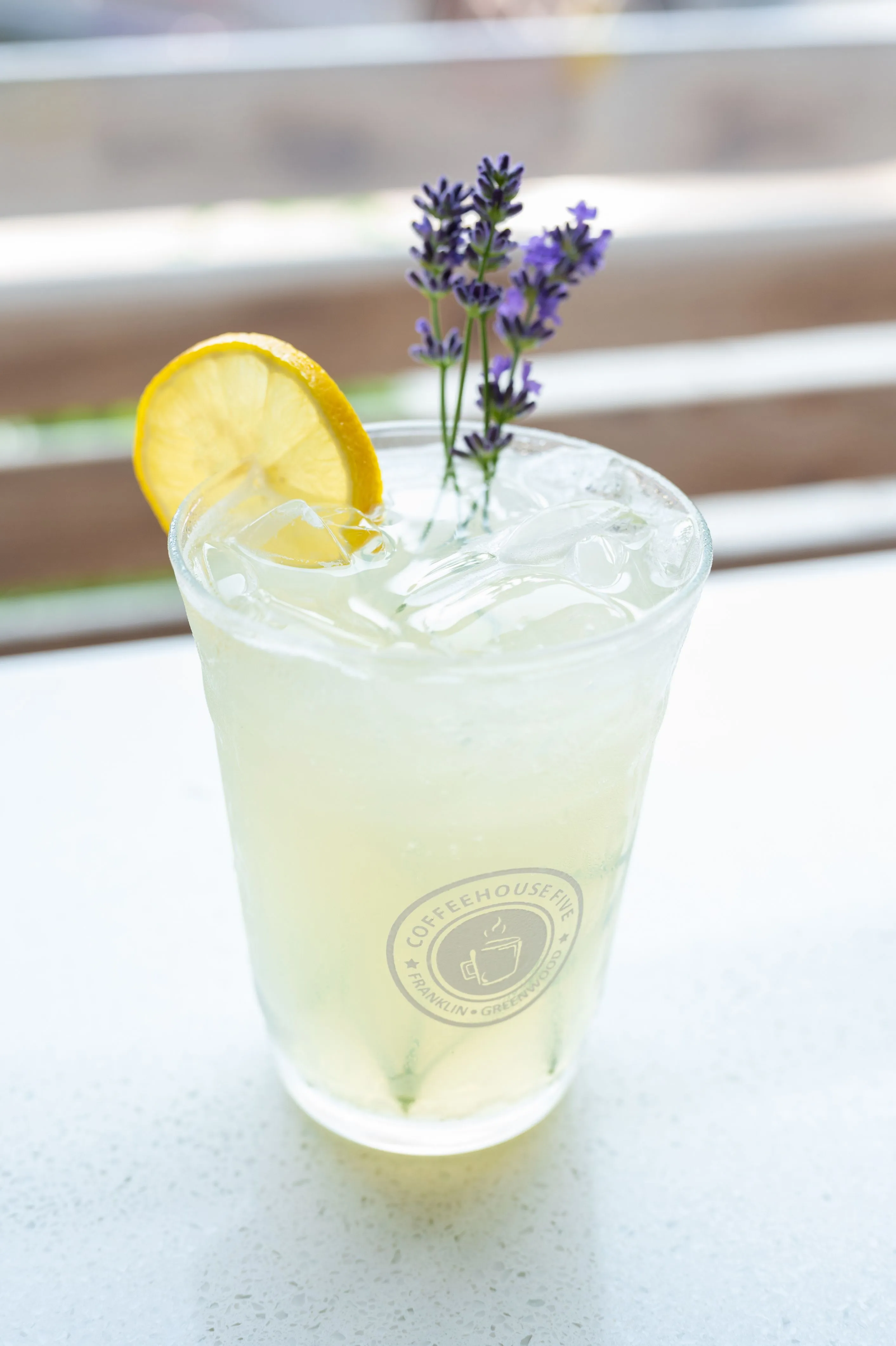 Iced lemonade garnished with a lemon slice and fresh lavender sprig in a glass imprinted with a coffeehouse logo.
