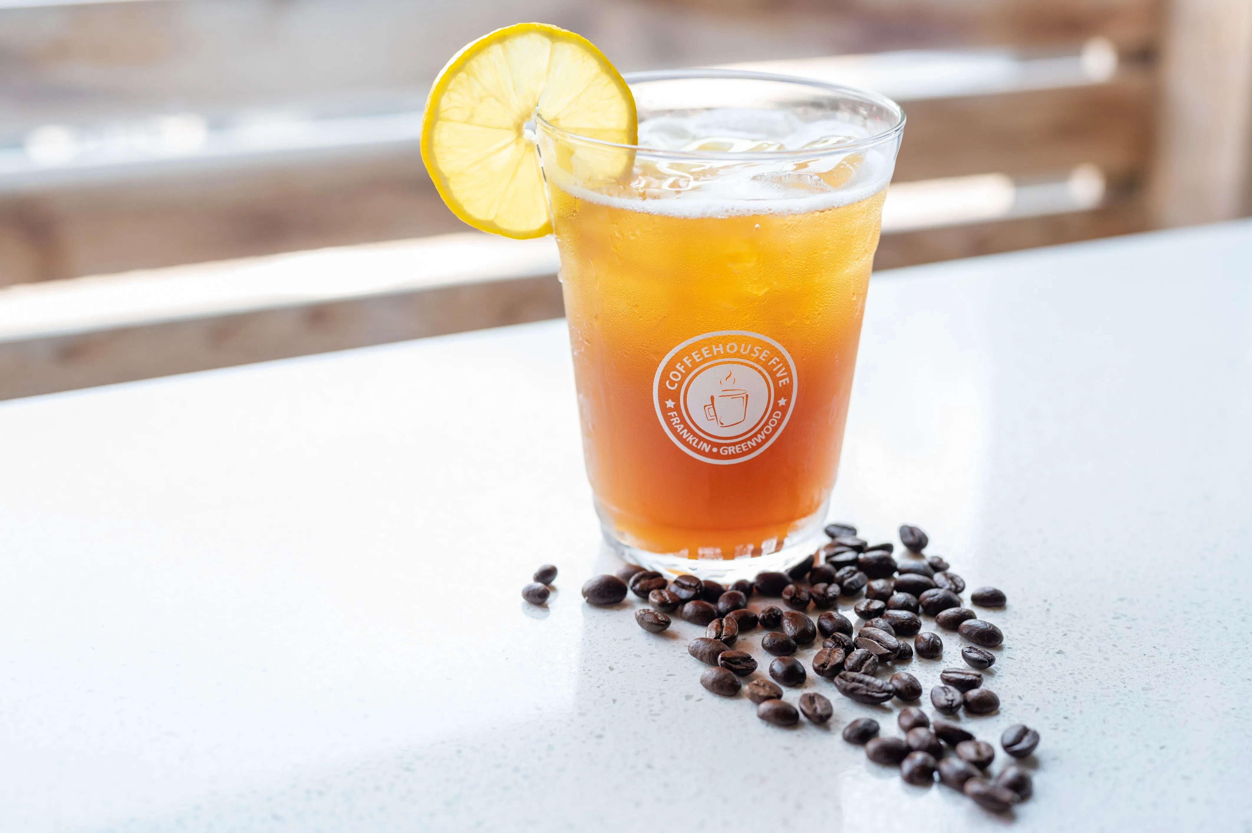 Iced coffee in a branded glass garnished with a lemon slice, surrounded by coffee beans on a table.