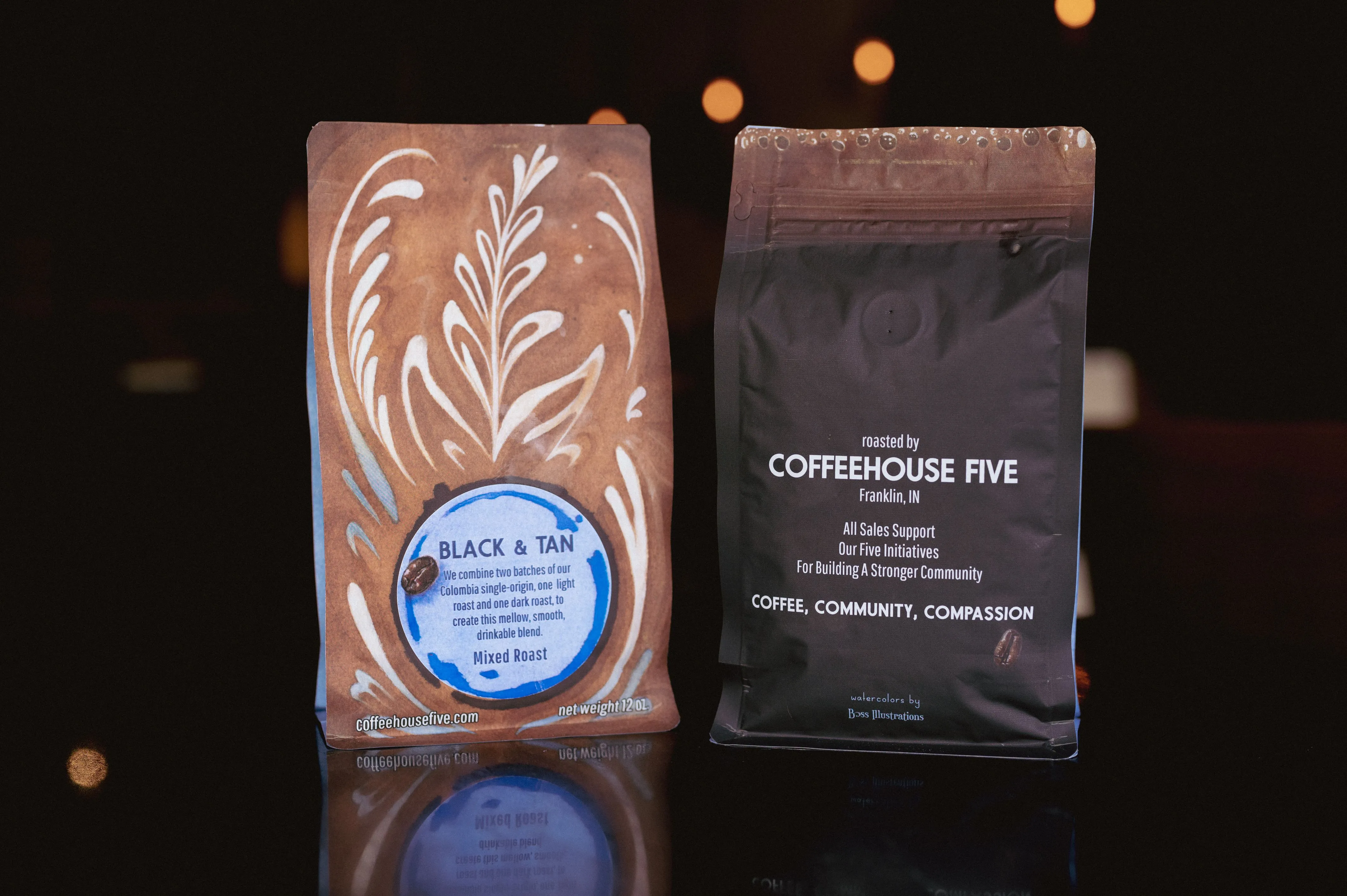 Two bags of Coffeehouse Five coffee on a reflective surface, one labeled 'Black & Tan' with a light brown design, and the other black with brand details and the words 'Coffee, Community, Compassion'.
