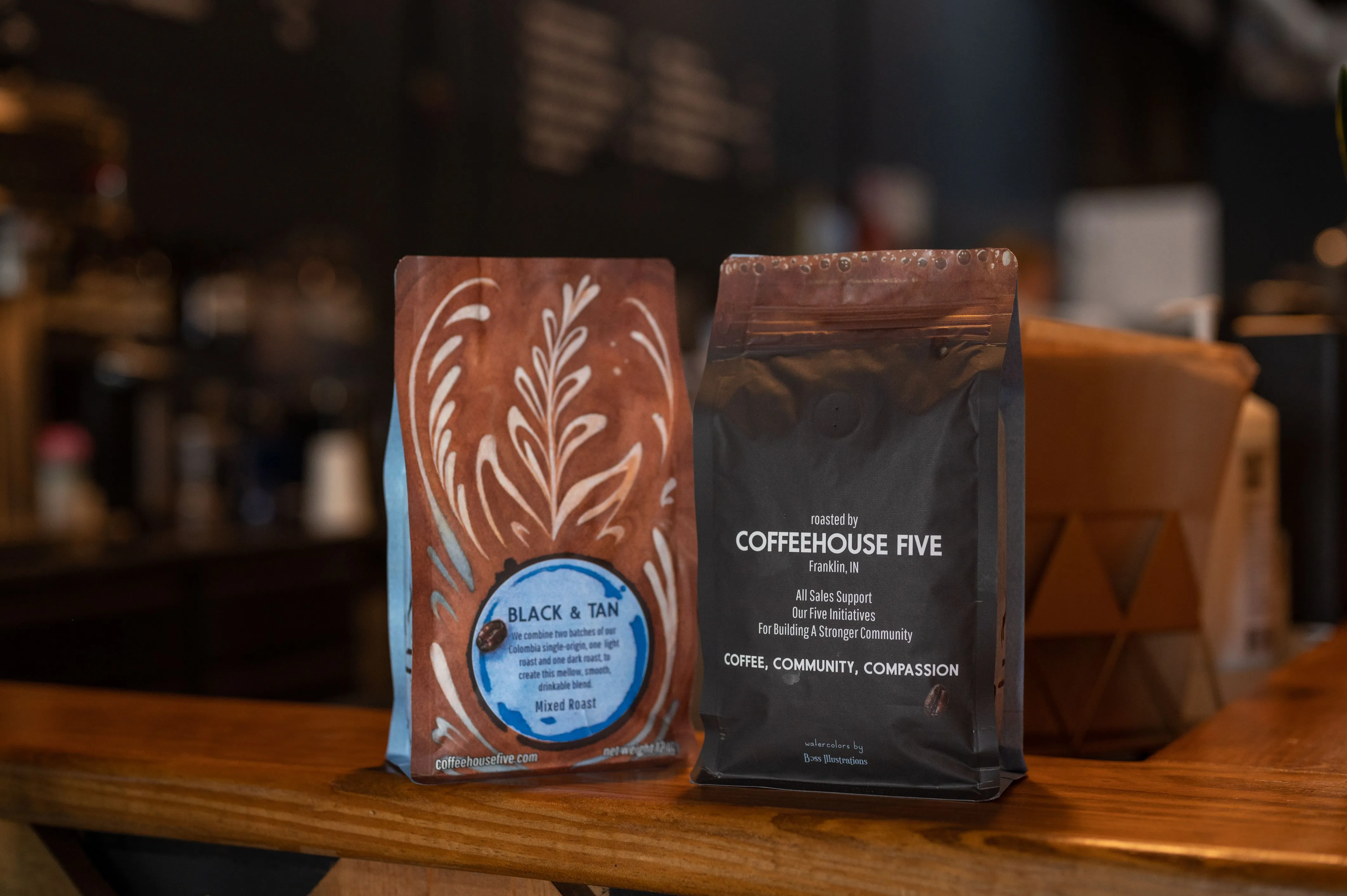 Two bags of Coffeehouse Five brand coffee on a wooden counter with a blurred coffee shop background.
