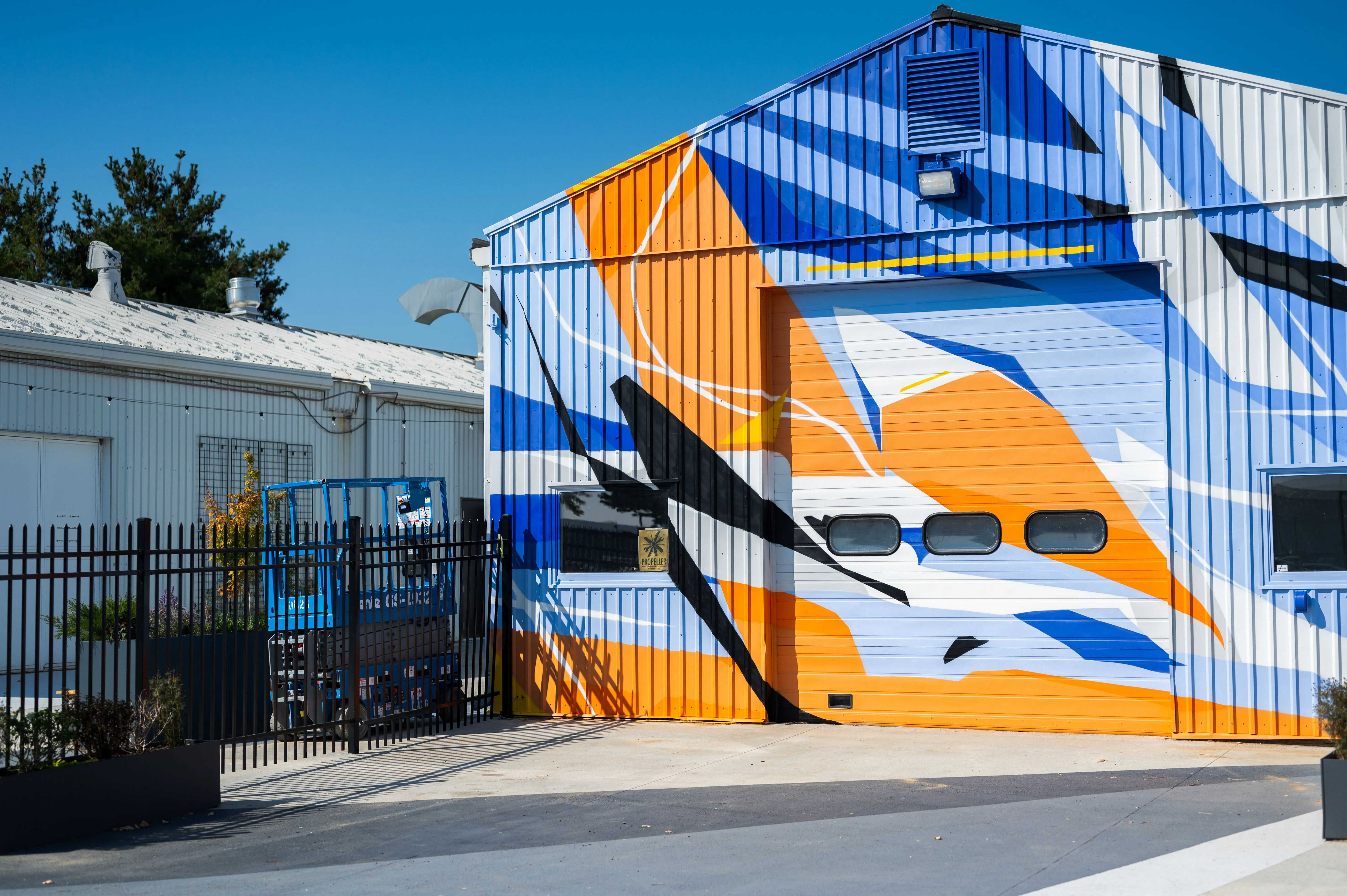 Industrial building with colorful abstract mural painted on the facade, featuring geometric shapes and stripes in blue, orange, and white, with gated entryway and clear blue sky.