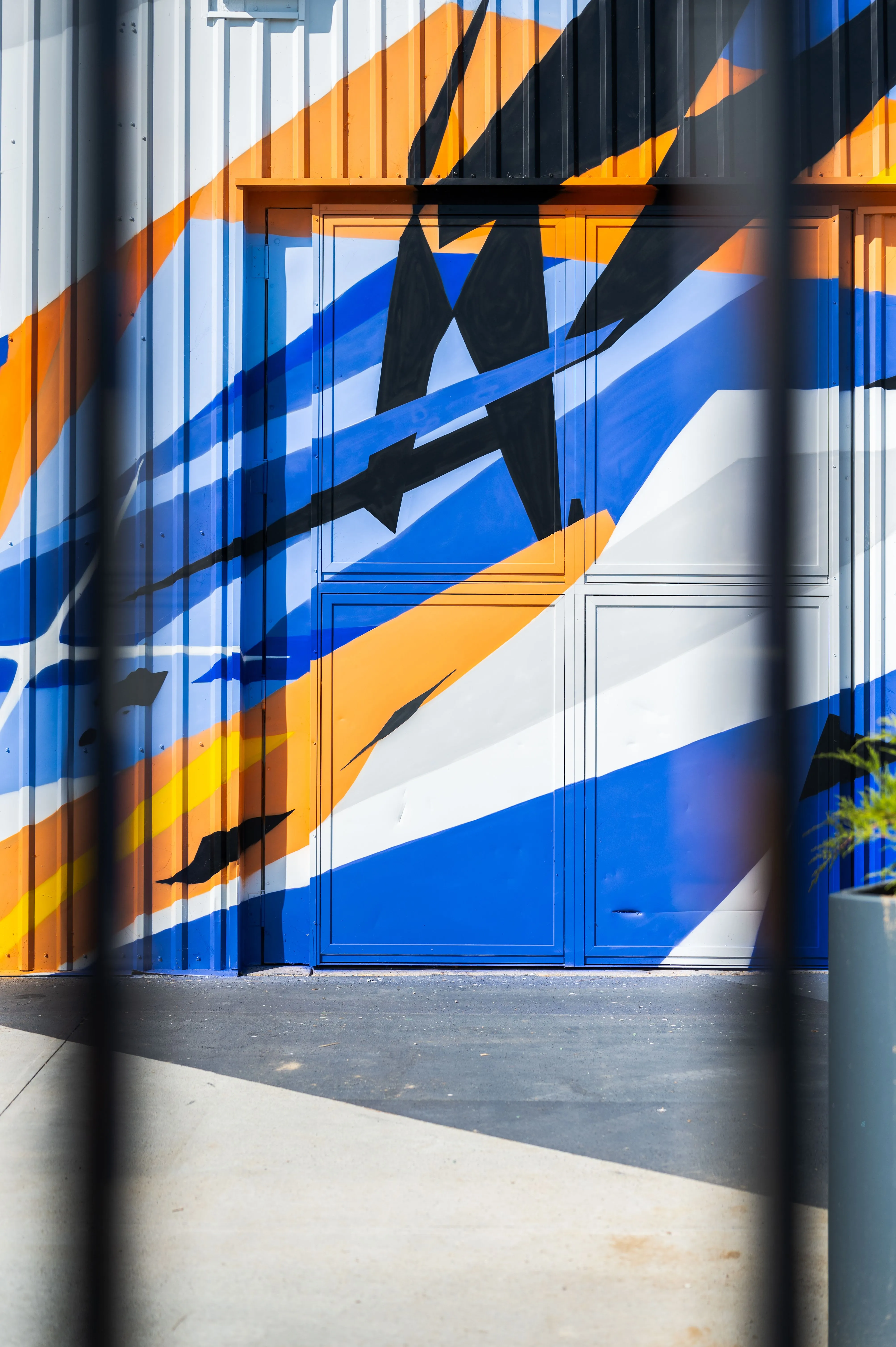 A colorful mural on the exterior of a building featuring geometric shapes in blue, white, and orange, with a blue door and shadows of nearby objects cast across the surface.