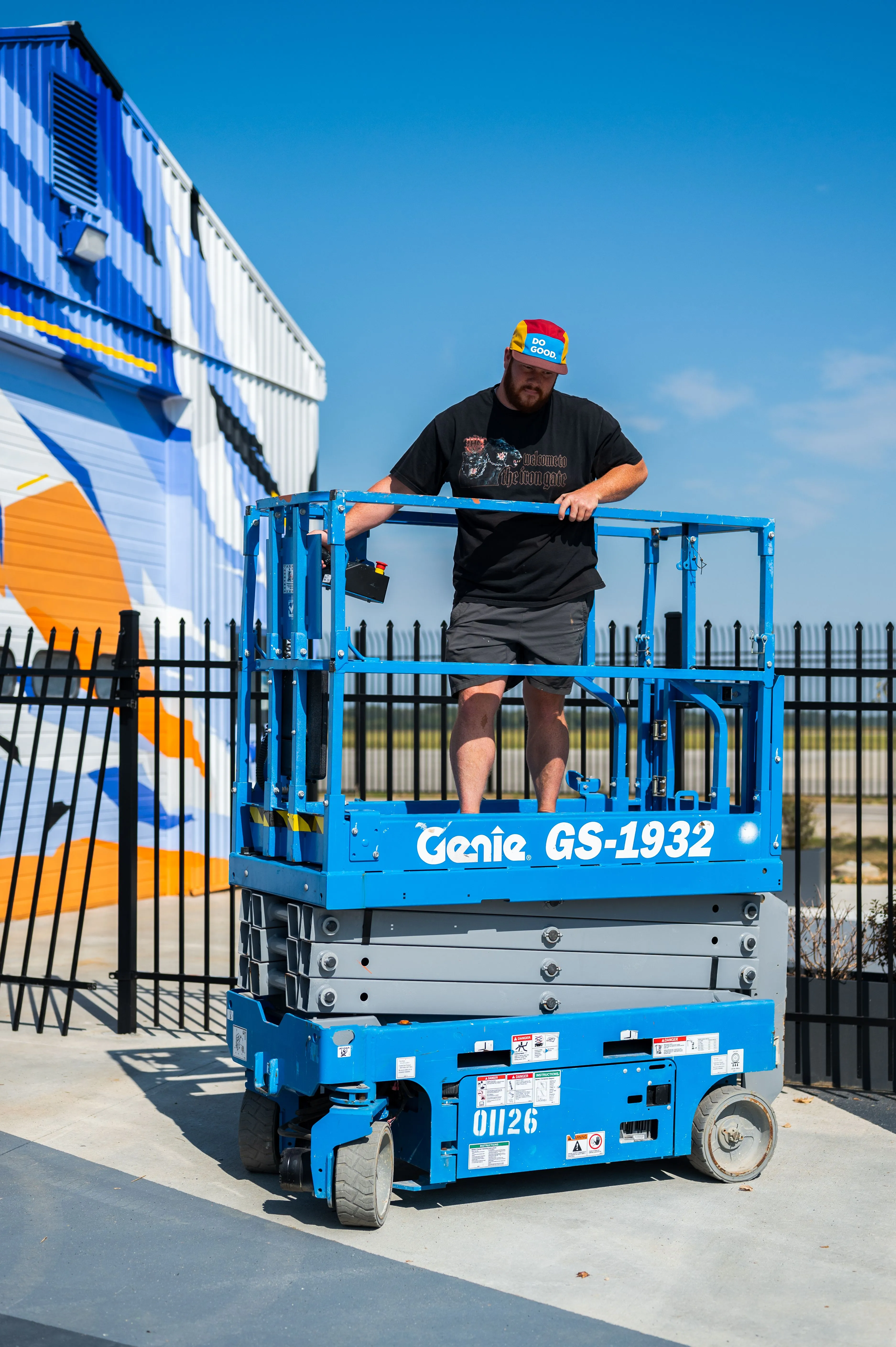 Man standing on a blue Genie GS-1932 scissor lift in front of an industrial building with blue and white striped walls.