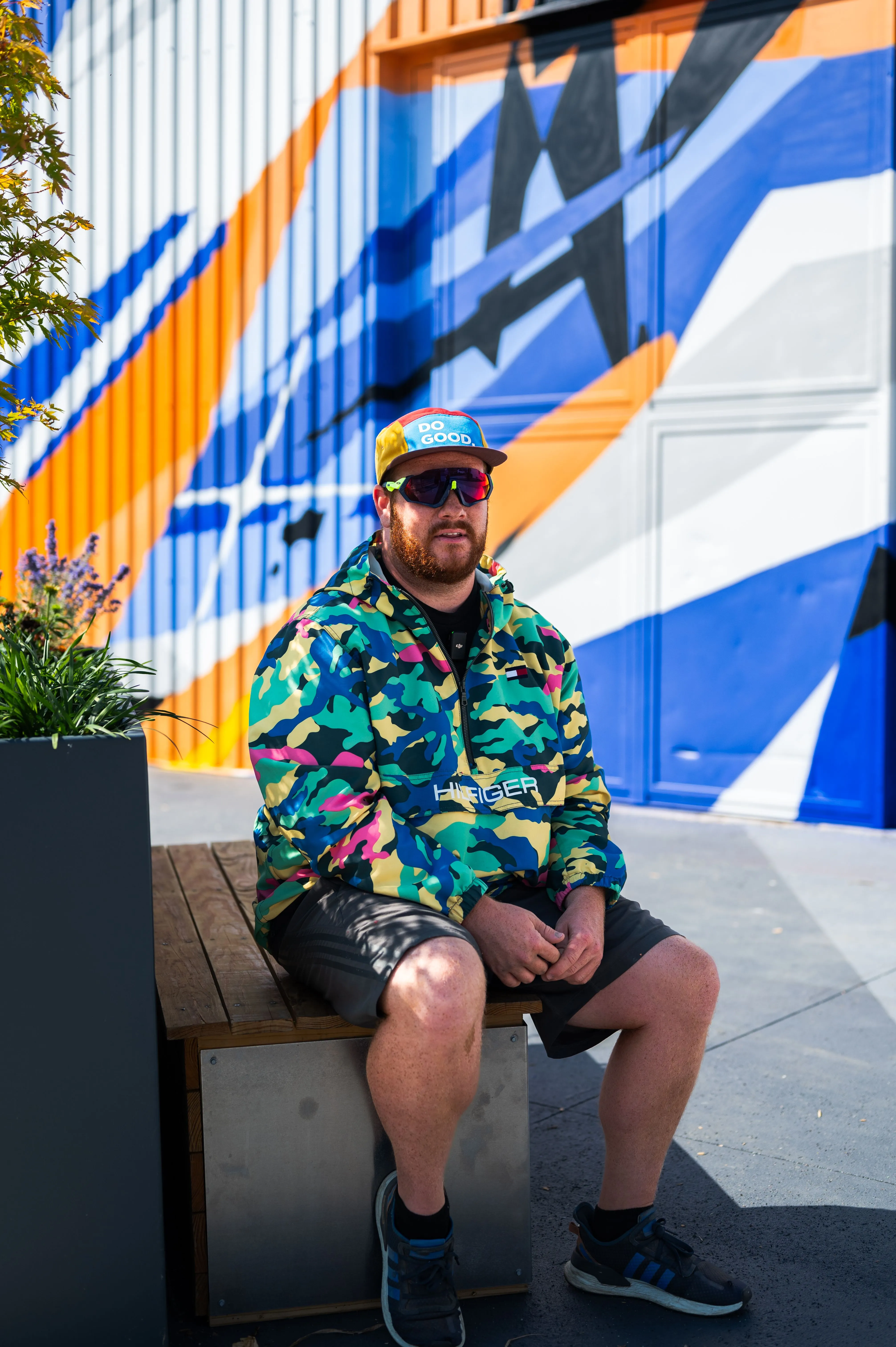 Man sitting on a bench wearing a colorful camouflaged jacket, sunglasses, and a cap against a vibrant geometric mural background.