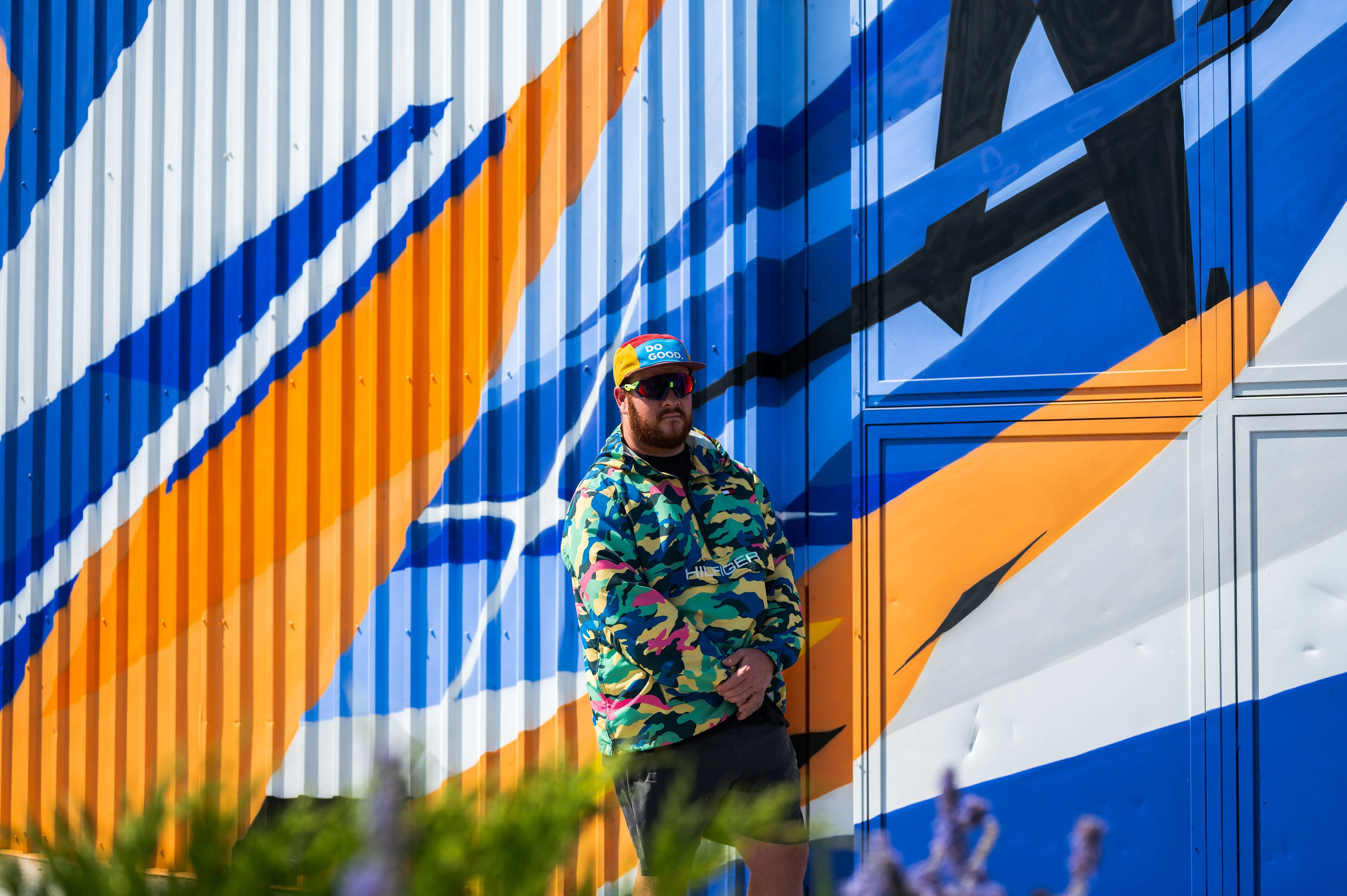Man standing in front of a colorful abstract mural, wearing a patterned jacket and sunglasses, with hands in pockets.