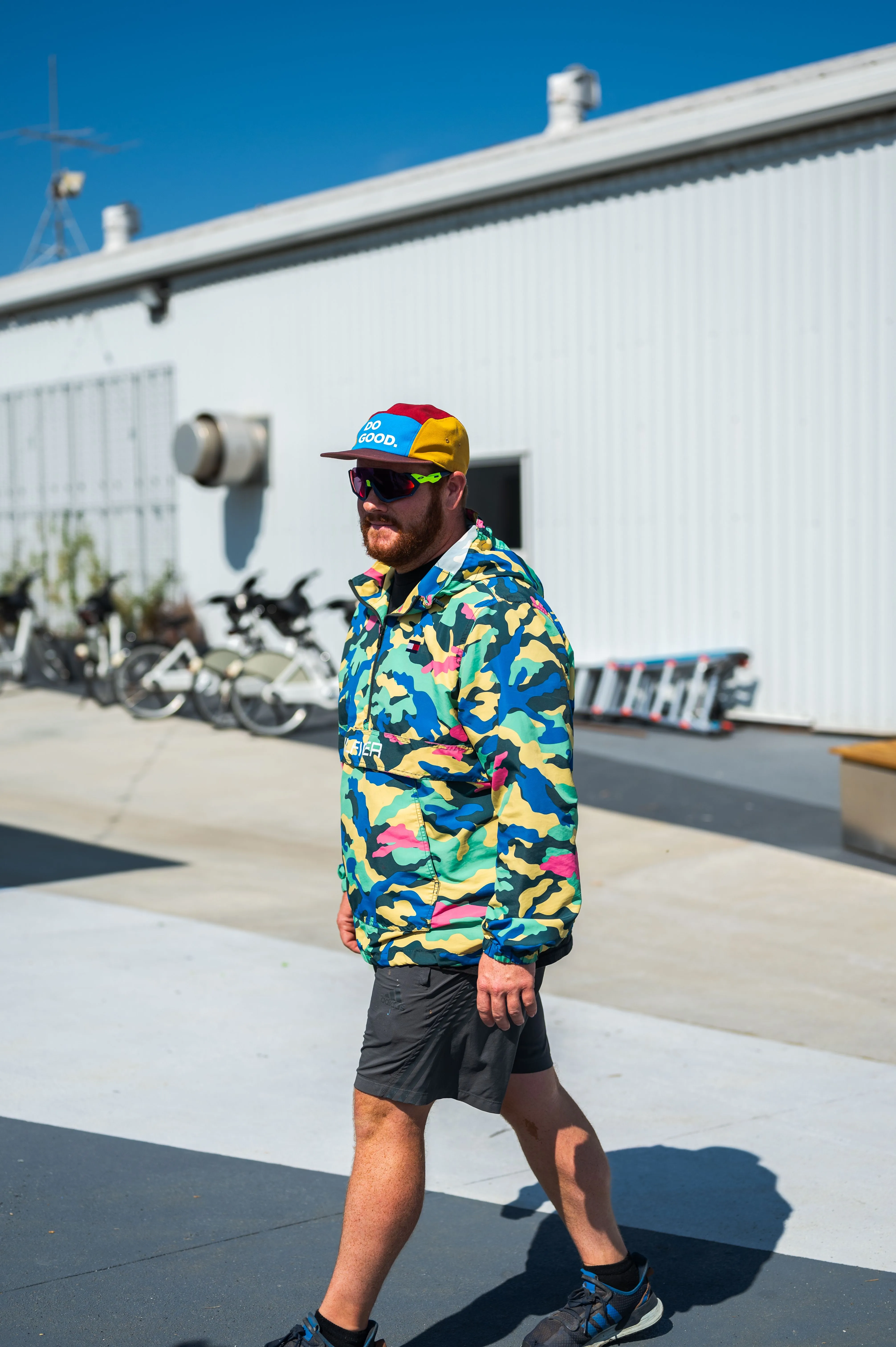 Man wearing a colorful camo jacket and multicolored cap walking in a sunny outdoor area with bicycles in the background.