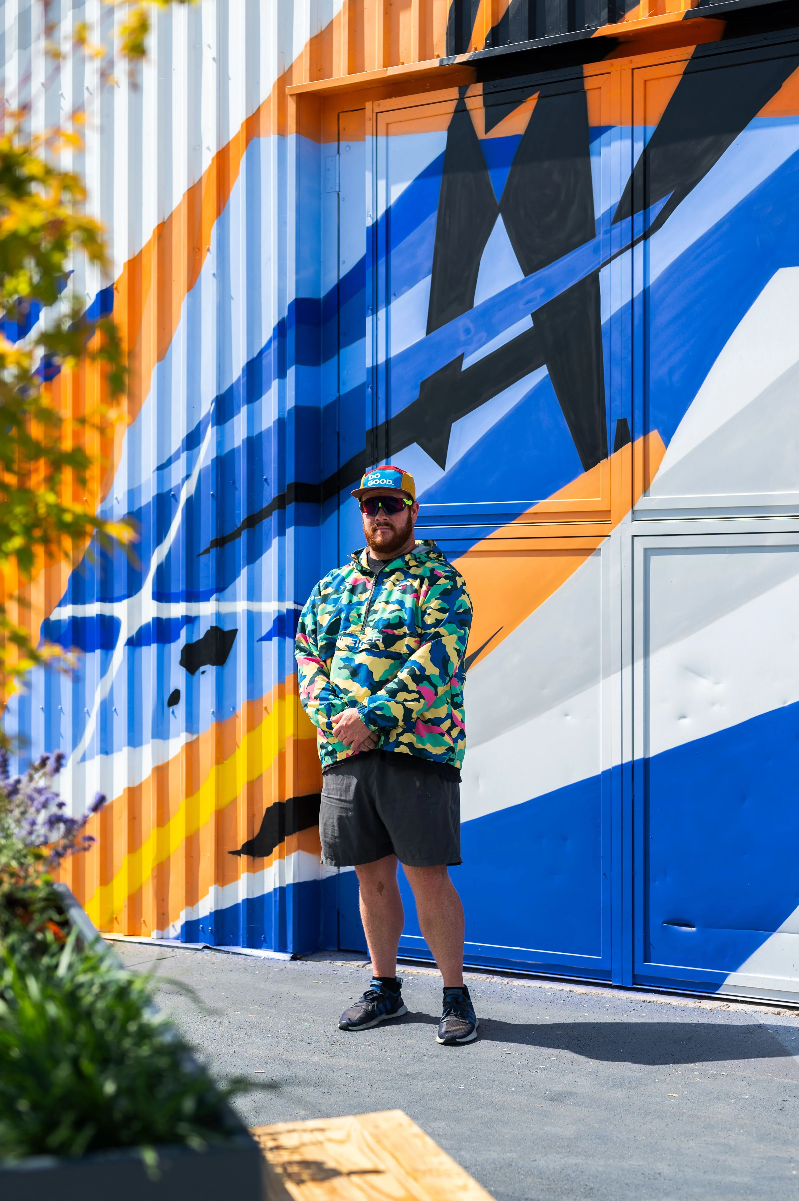 Man standing in front of a colorful container wall with an abstract design, wearing a camouflage jacket, shorts, sunglasses, and a cap.