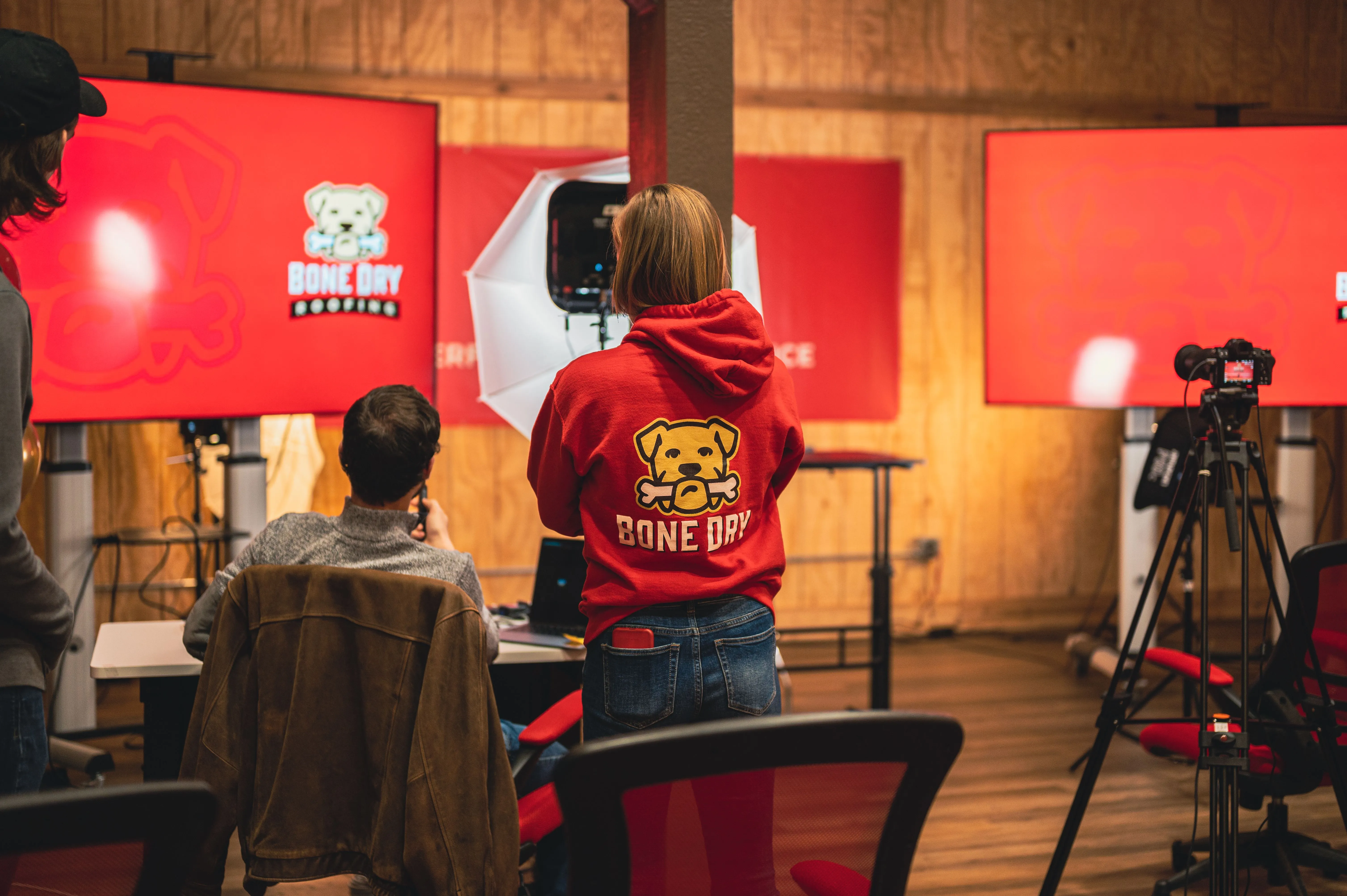 Individual in red hoodie watching over a video recording setup with multiple monitors and a camera on a tripod in an indoor setting.