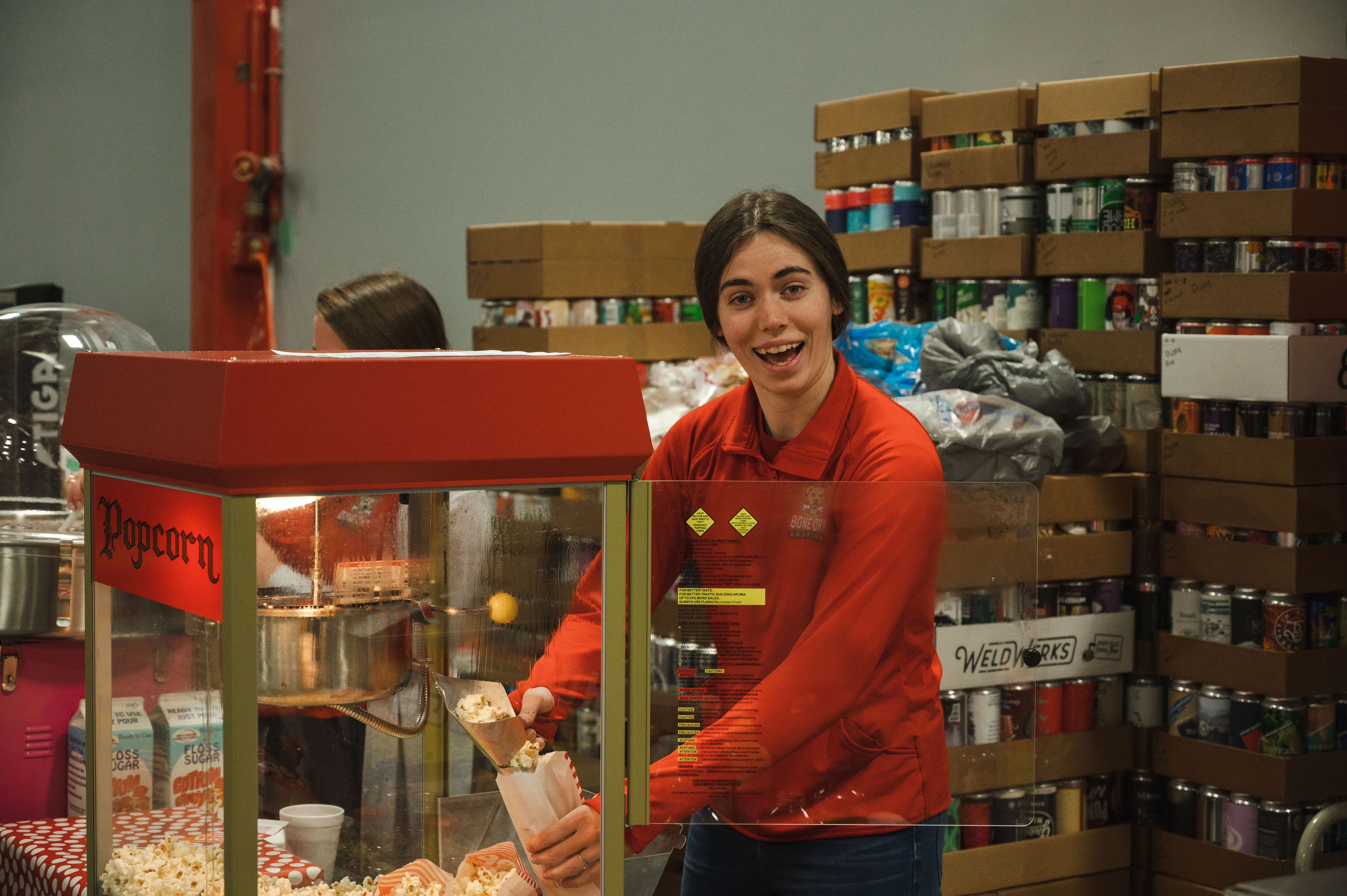 Worker smiling while standing behind a concession stand with popcorn and boxes in the background.
