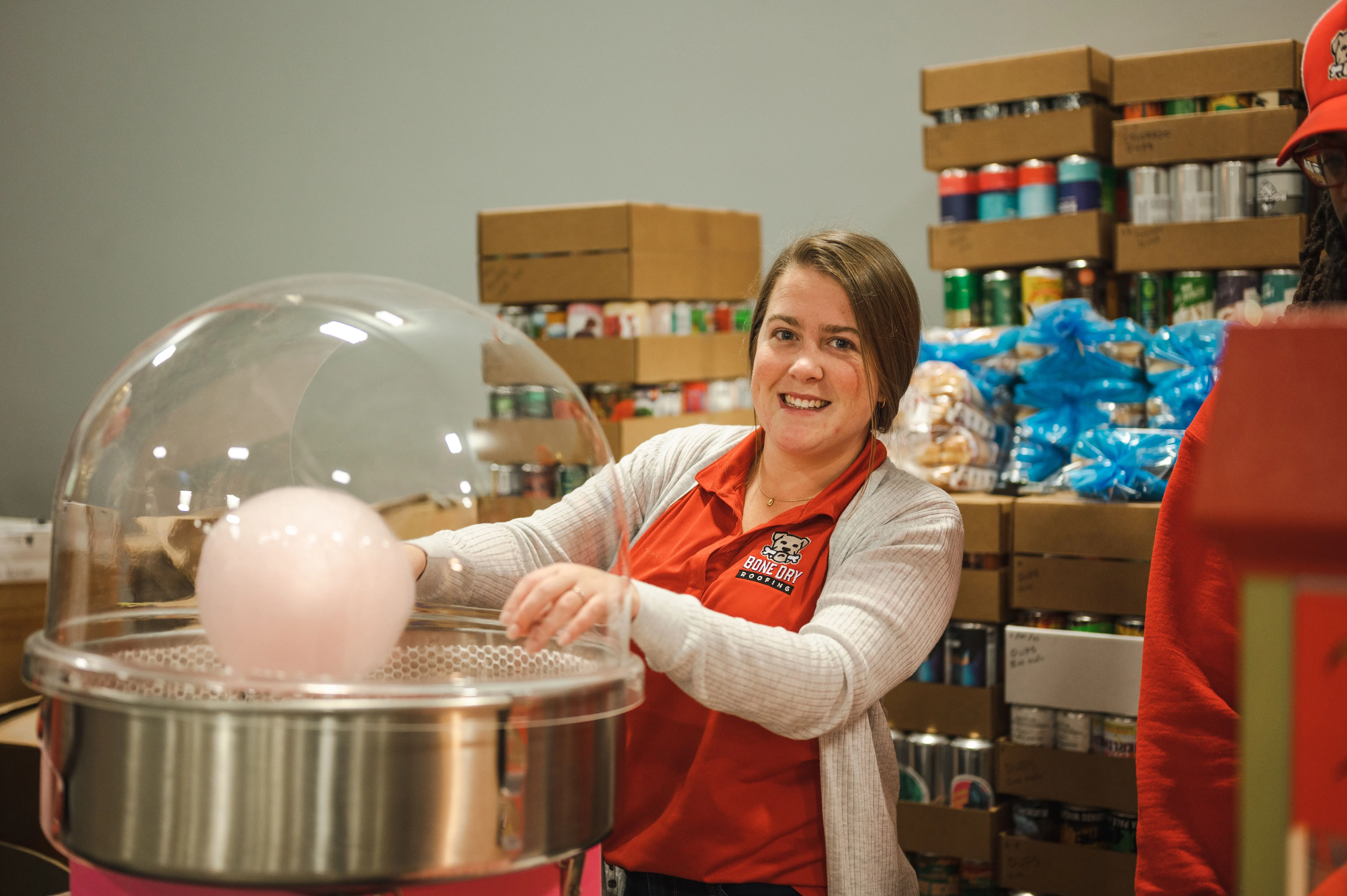 Smiling woman in a red apron making cotton candy at a concession stand with shelves of supplies in the background.