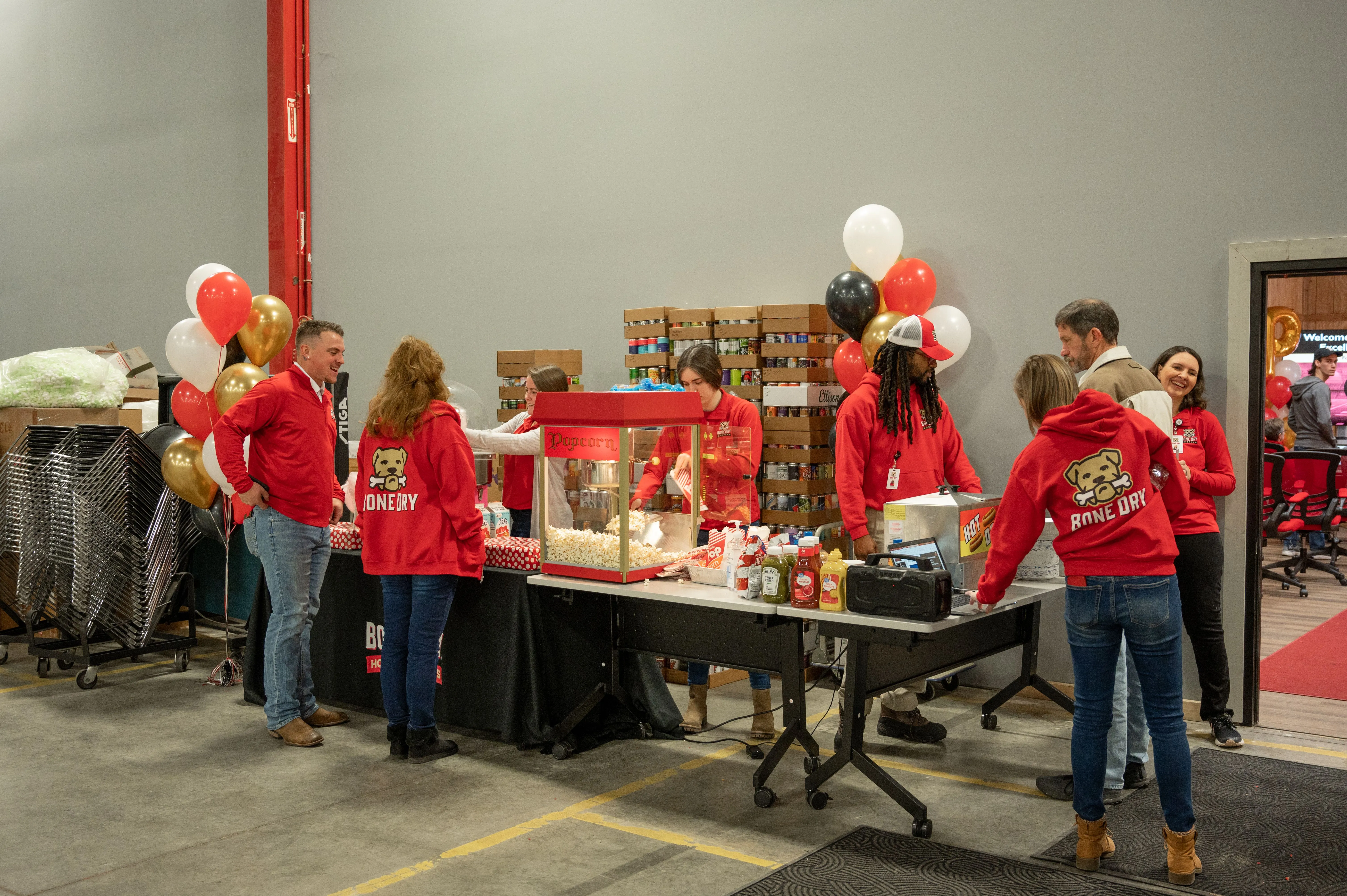 Group of people wearing red shirts gathered around a booth with red and white balloons at an indoor event.