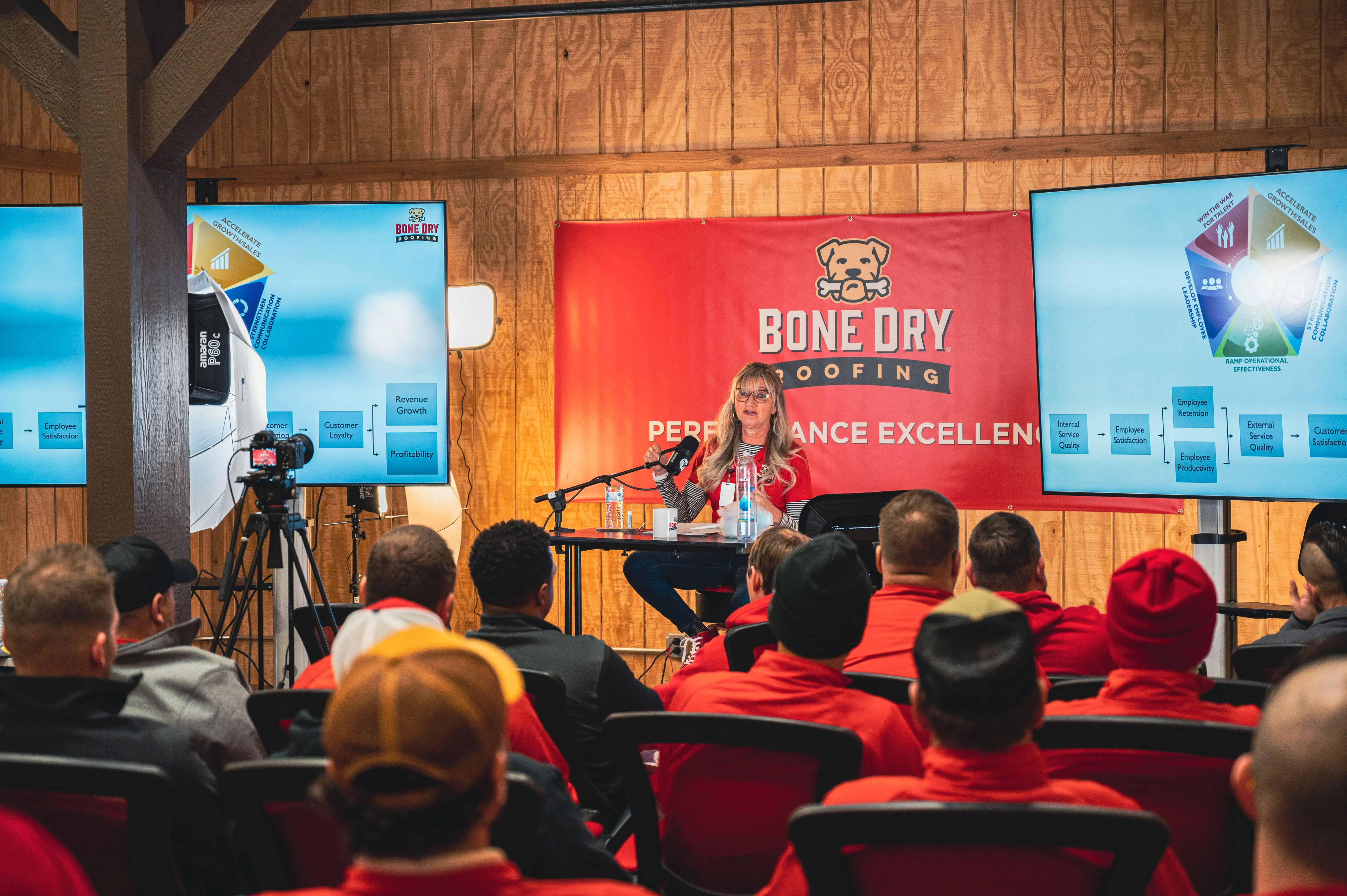 Woman presenting at a conference with attendees in red hats, video cameras recording, and banners reading "Bone Dry Roofing Company".