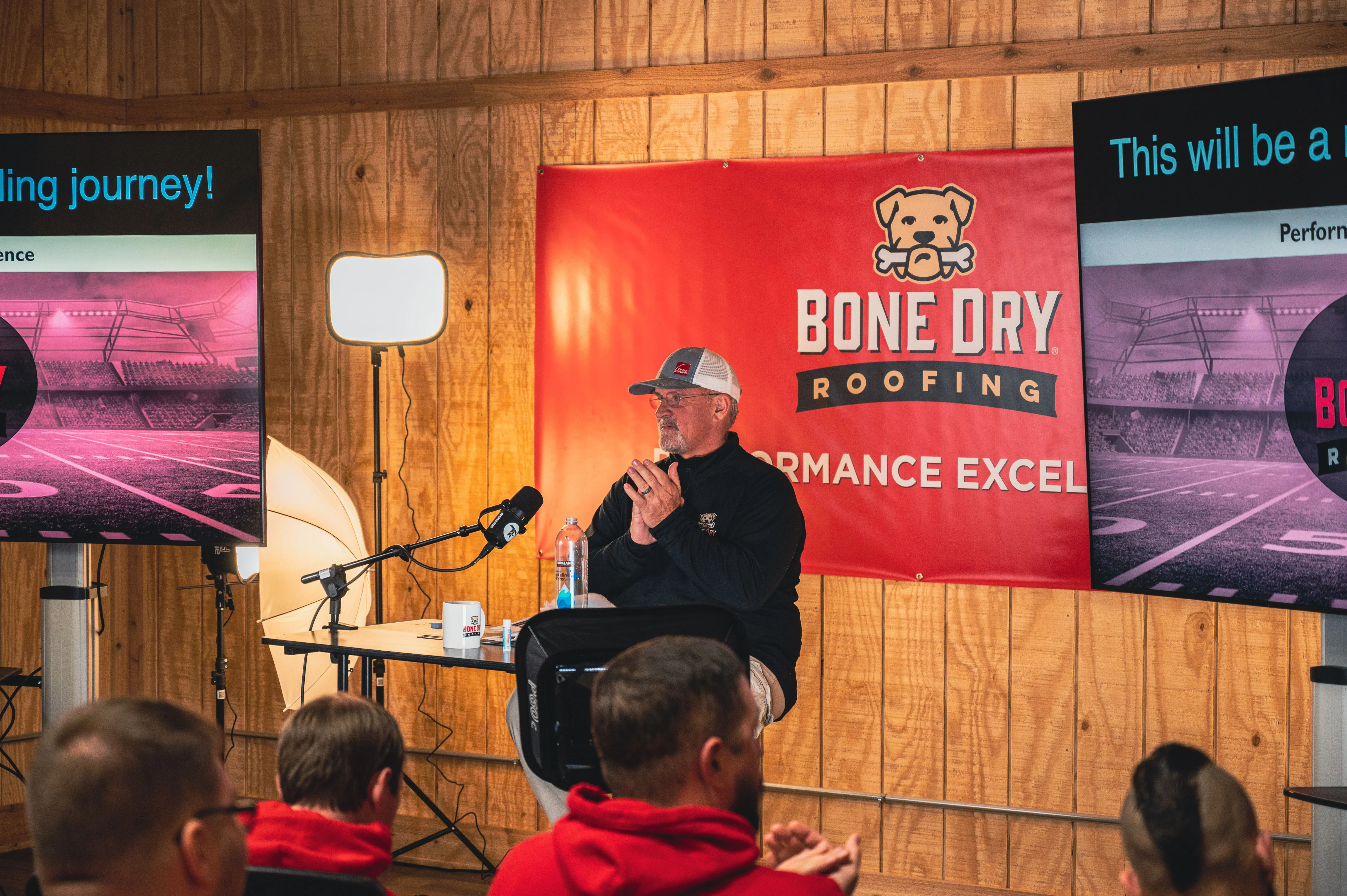 A speaker presenting to an audience with "Bone Dry Roofing Company" banners in the background.