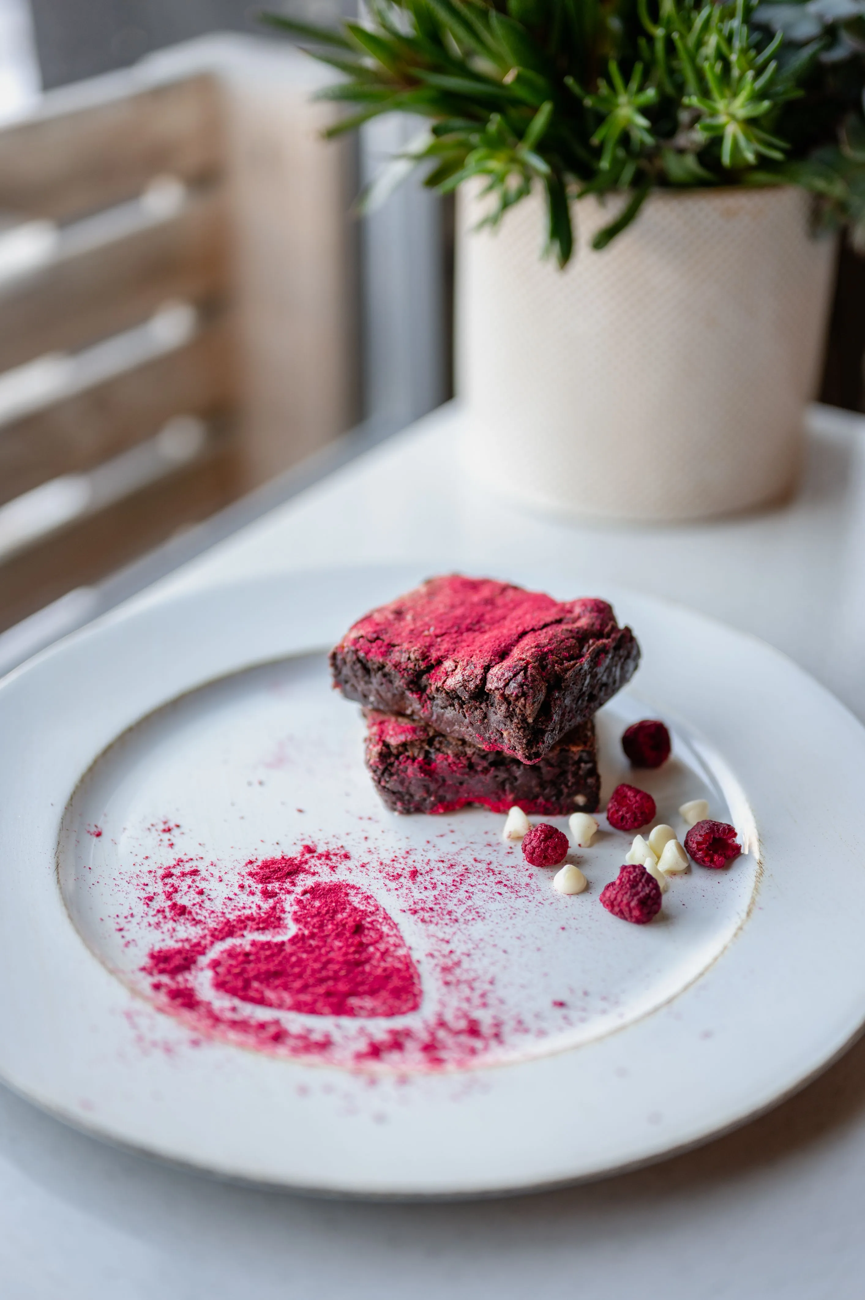 Gourmet presentation of a raspberry brownie on a white plate with freeze-dried raspberry powder shaped as a heart and scattered berries.