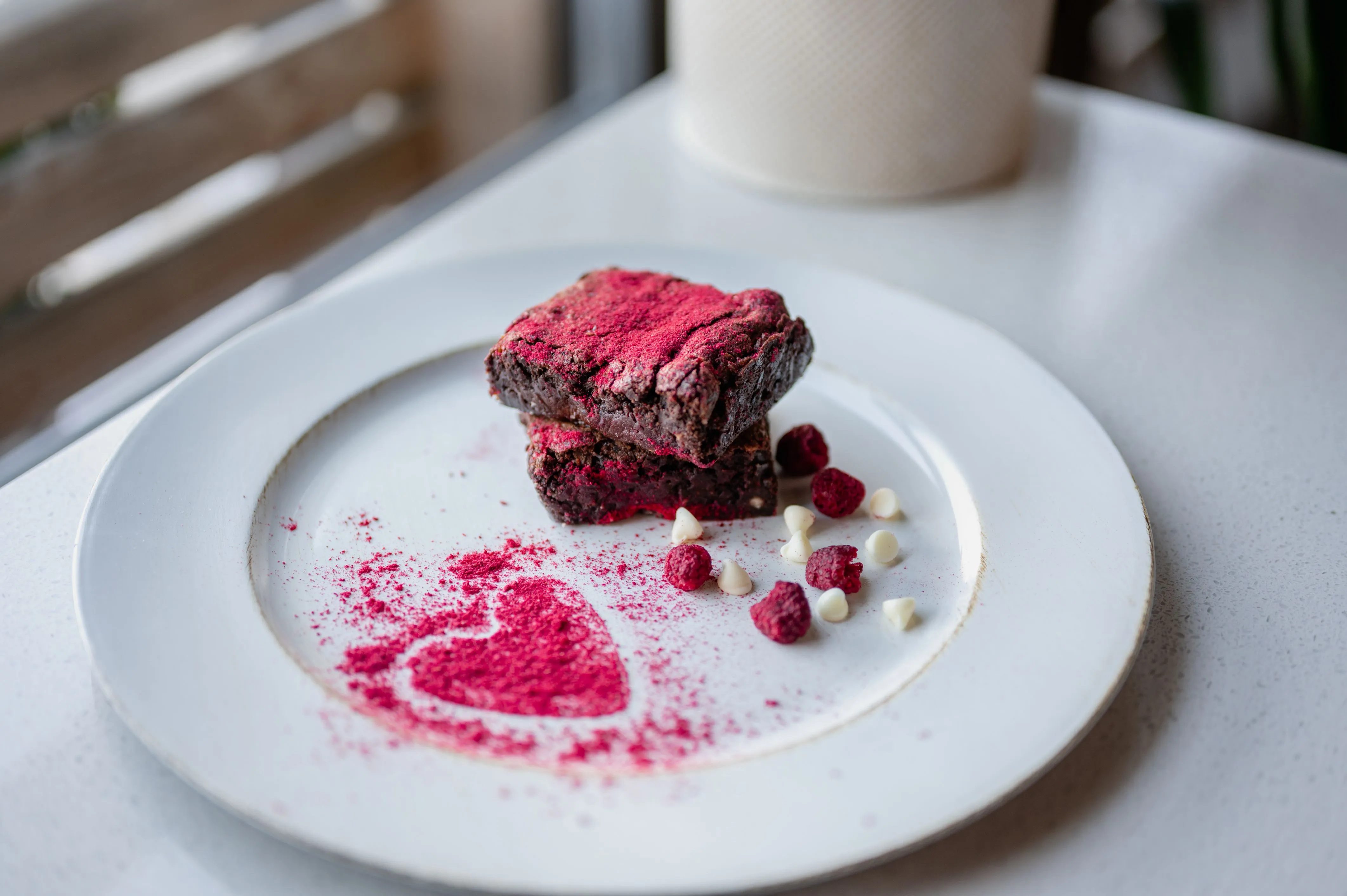 Red velvet brownies presented on a plate with freeze-dried raspberries and white chocolate chips, with a powdered heart-shaped decoration.