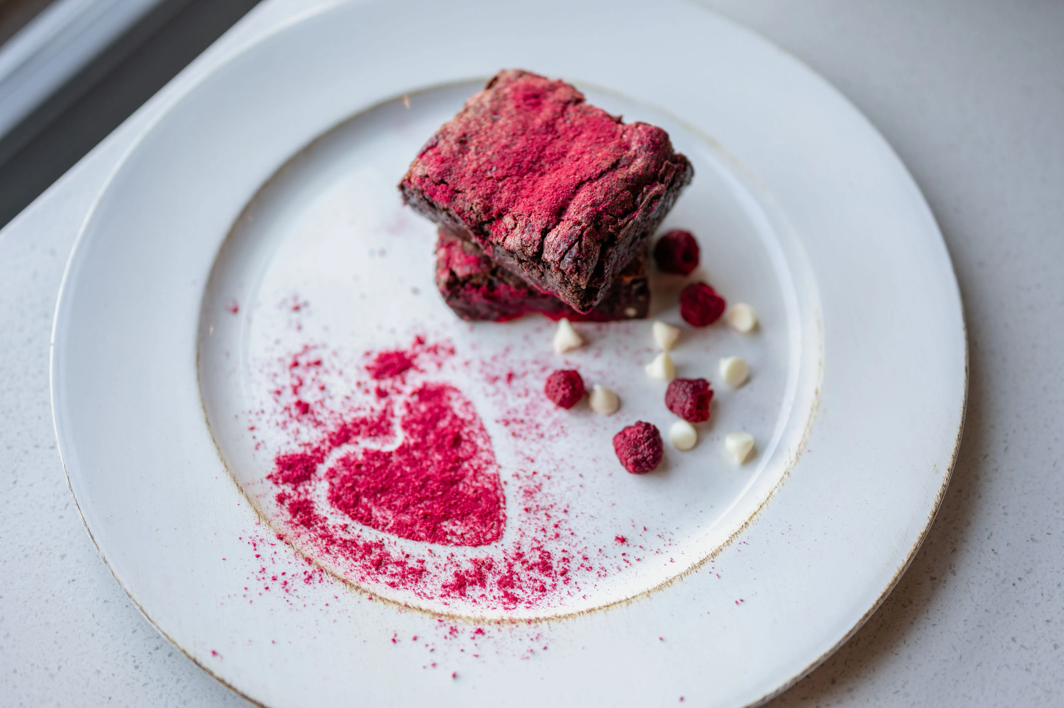 Gourmet brownie dusted with red powder on a white plate, adorned with a decorative powdered sugar heart and scattered raspberries and white chocolate drops.