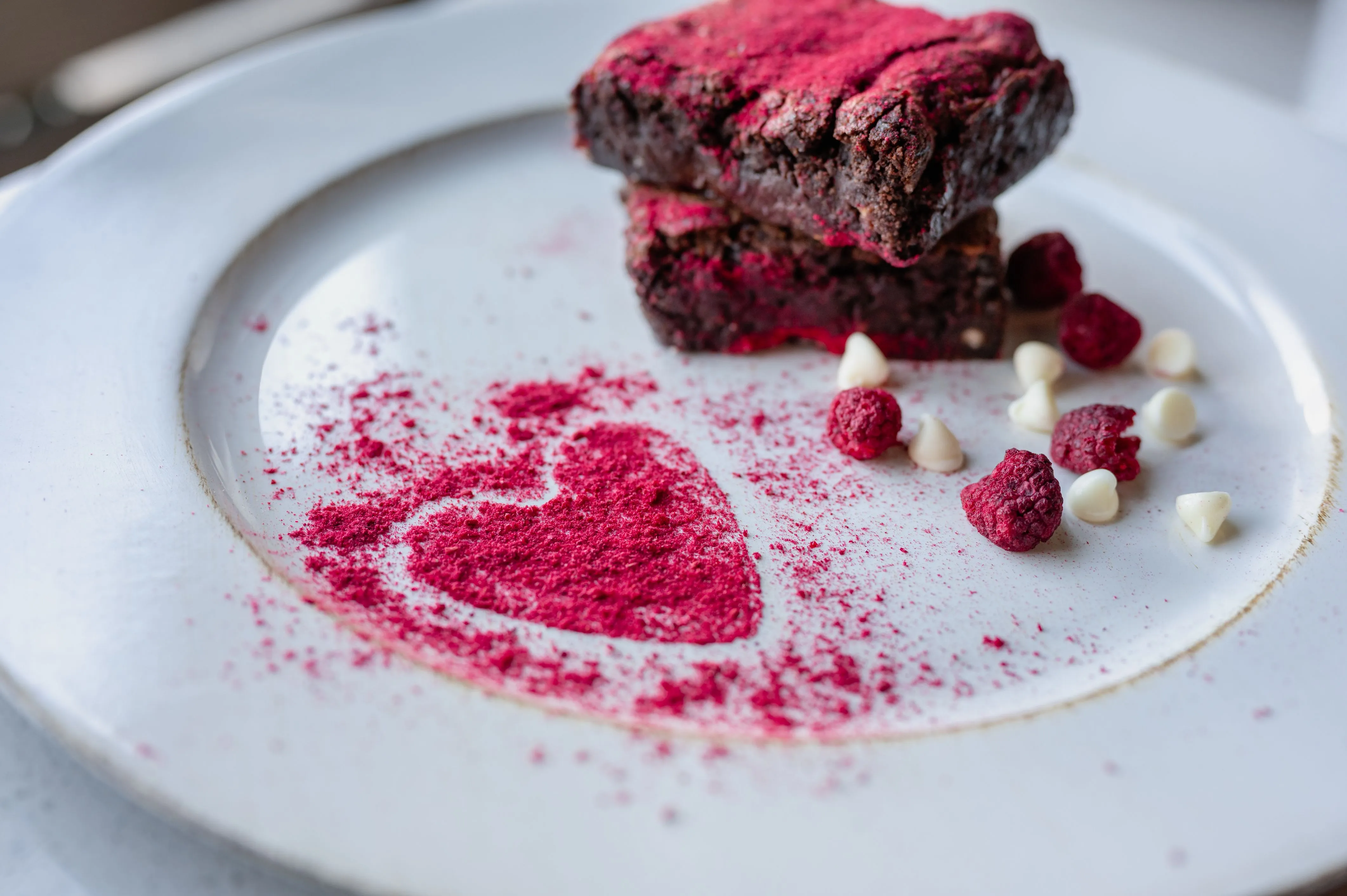 A gourmet brownie dessert elegantly presented on a white plate with a heart-shaped raspberry dusting and garnished with freeze-dried raspberries and white chocolate chips.