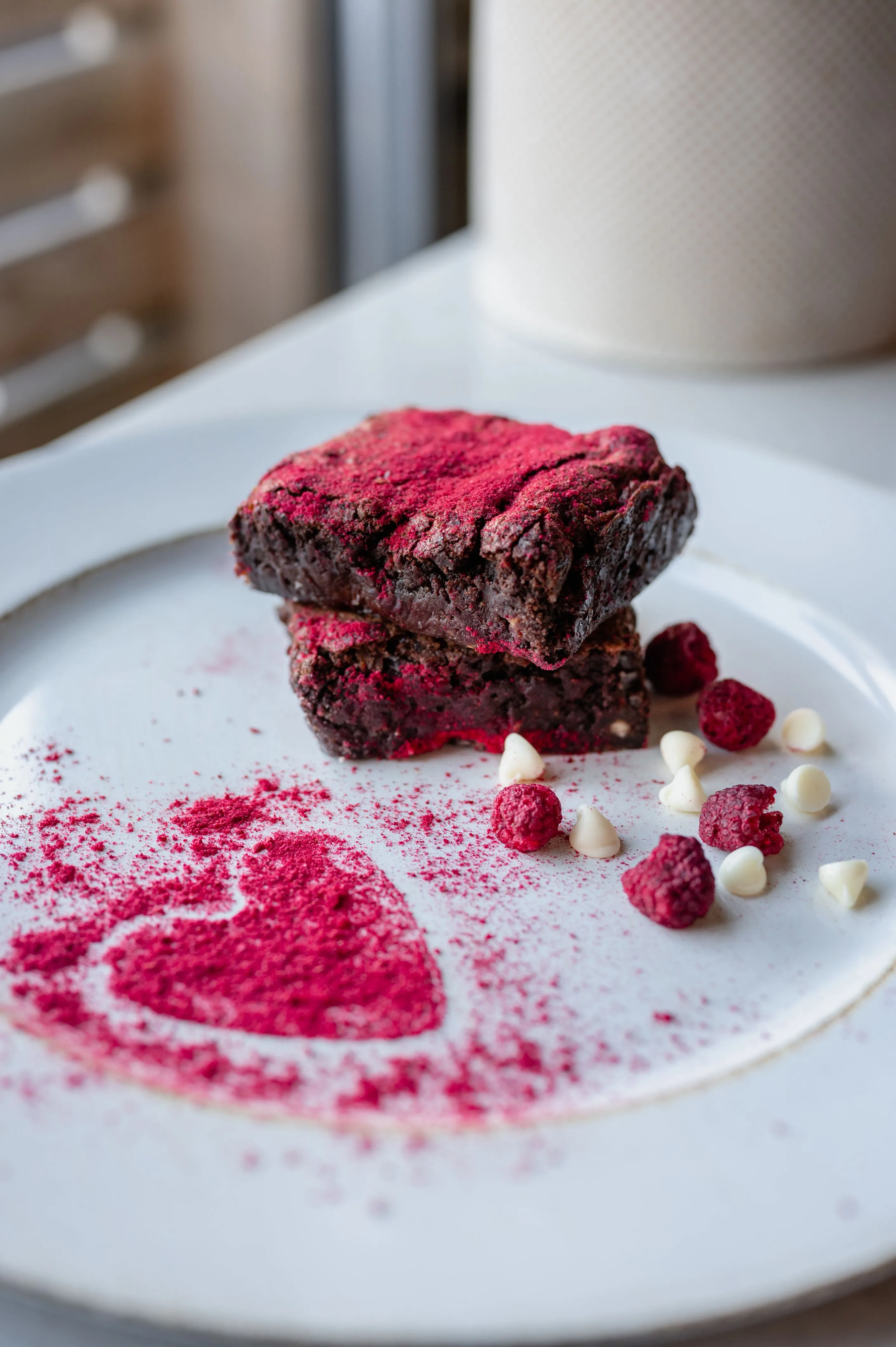 Two chocolate brownies dusted with red powder, served on a plate with a heart-shaped pattern and garnished with white chocolate drops and freeze-dried raspberries.