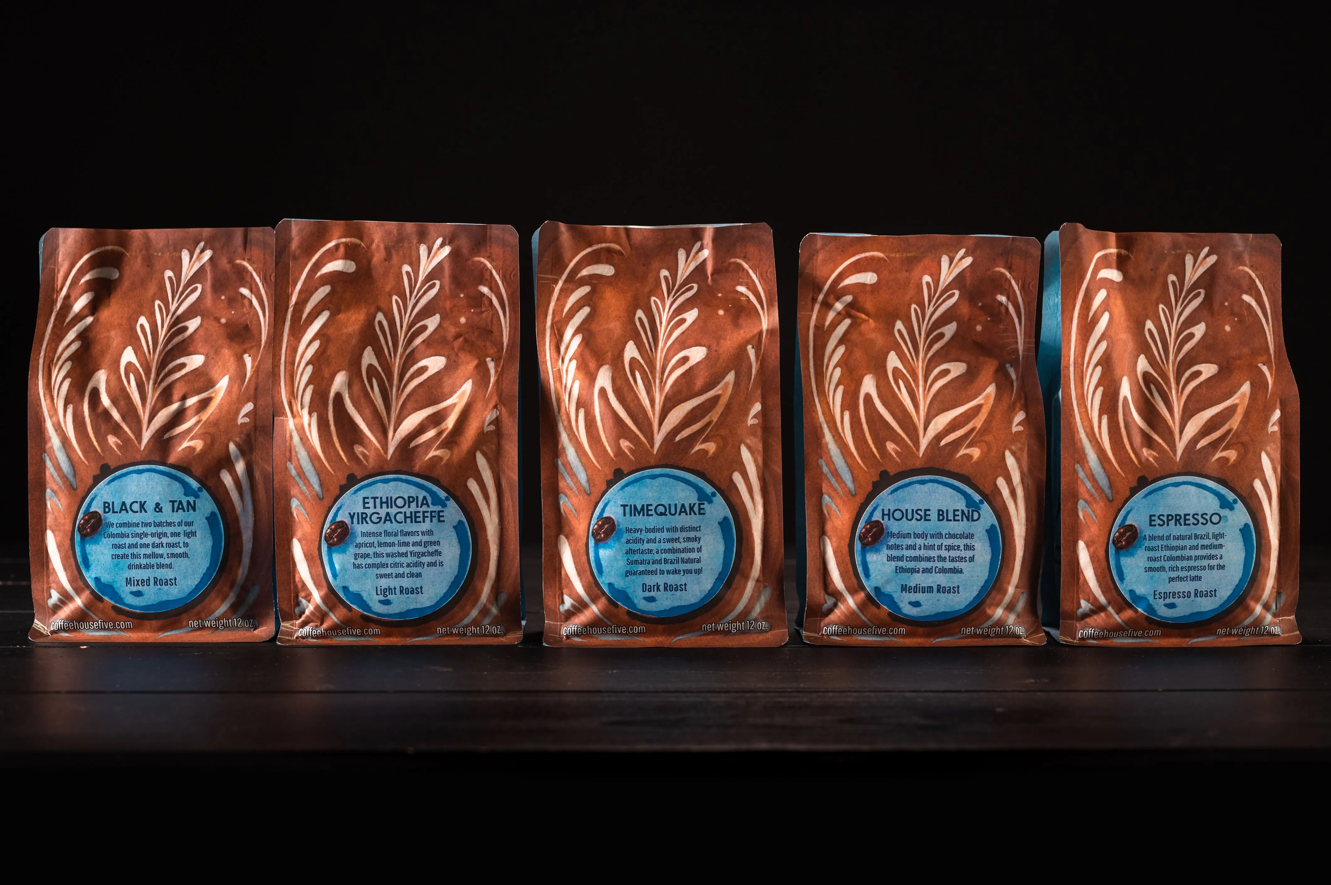 Five bags of artisanal coffee on a shelf, each with a unique blend and roast type, featuring decorative leaf patterns on a dark background.