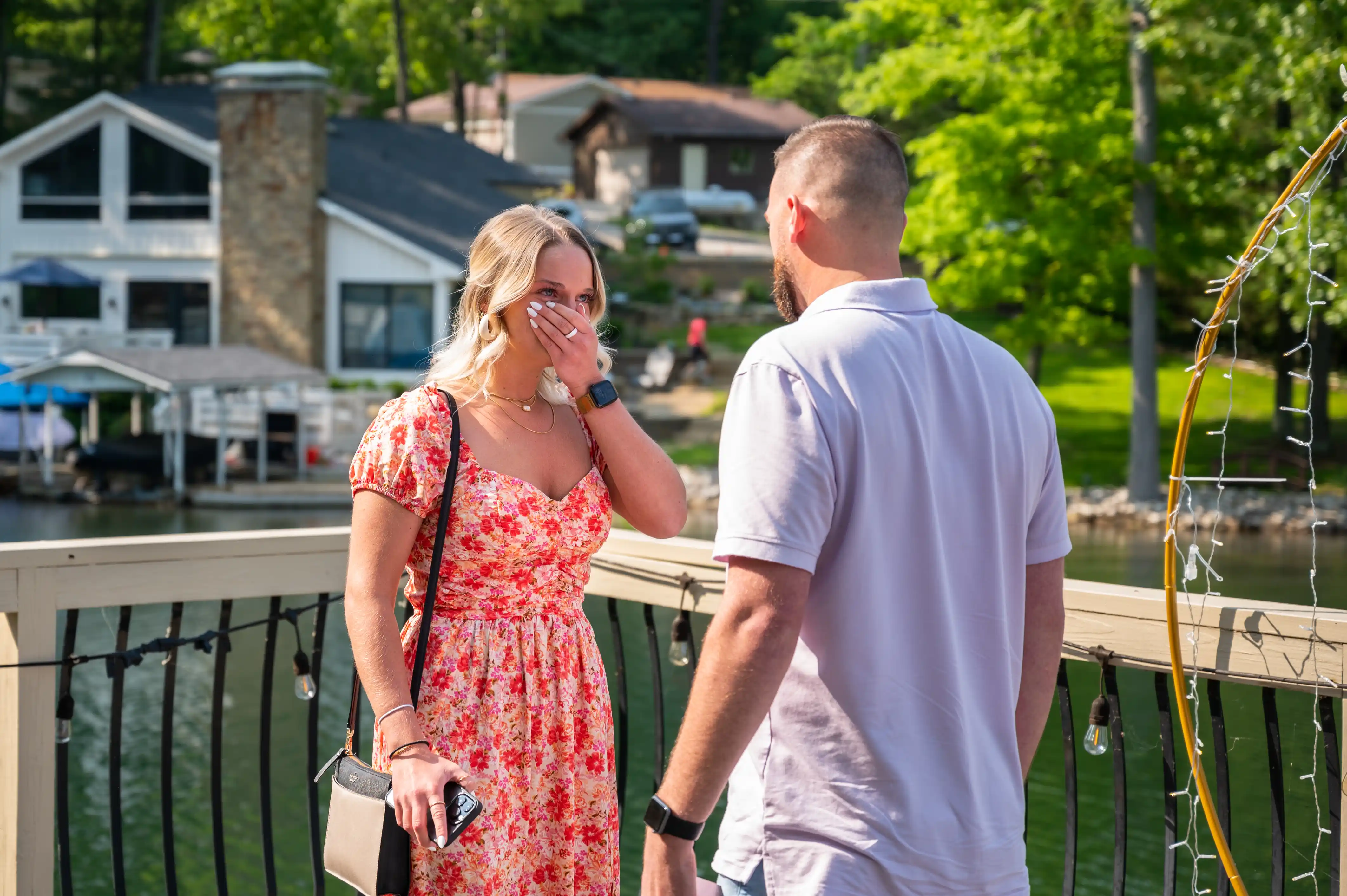 Two people having a conversation by a lakeside railing with houses and trees in the background.