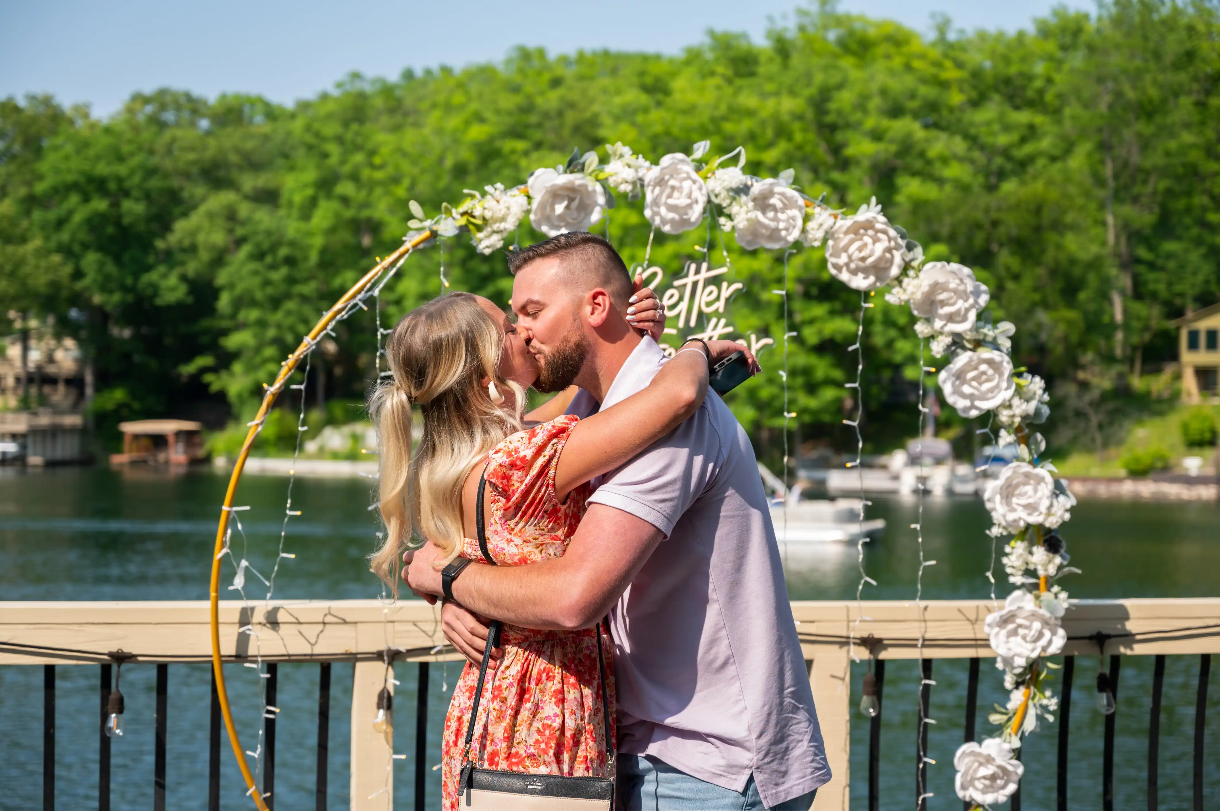 Couple embracing and kissing under a floral arch on a bridge with a lake and boats in the background