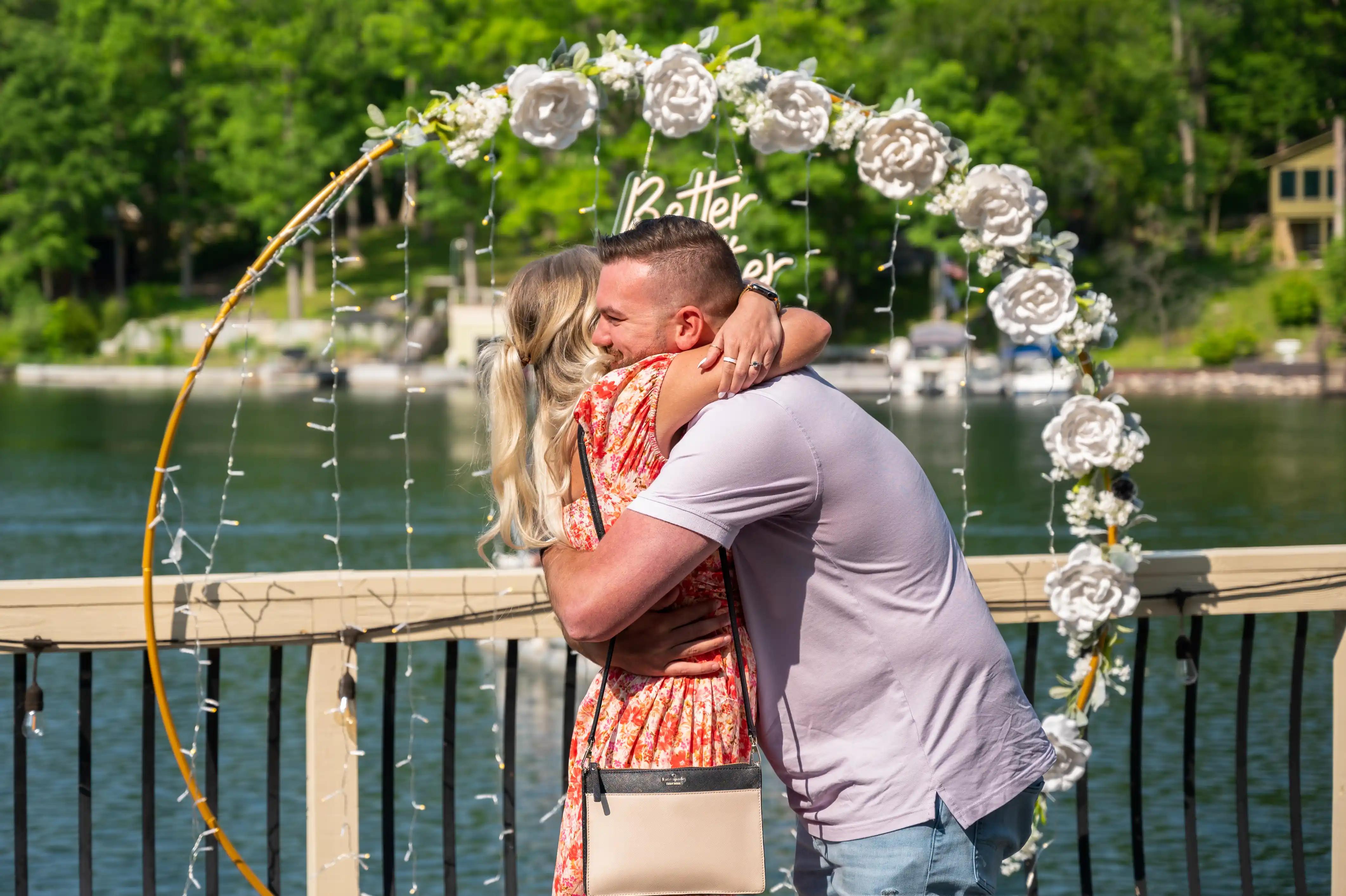 Couple embracing under a floral arch by a lakeside with boats in the background.