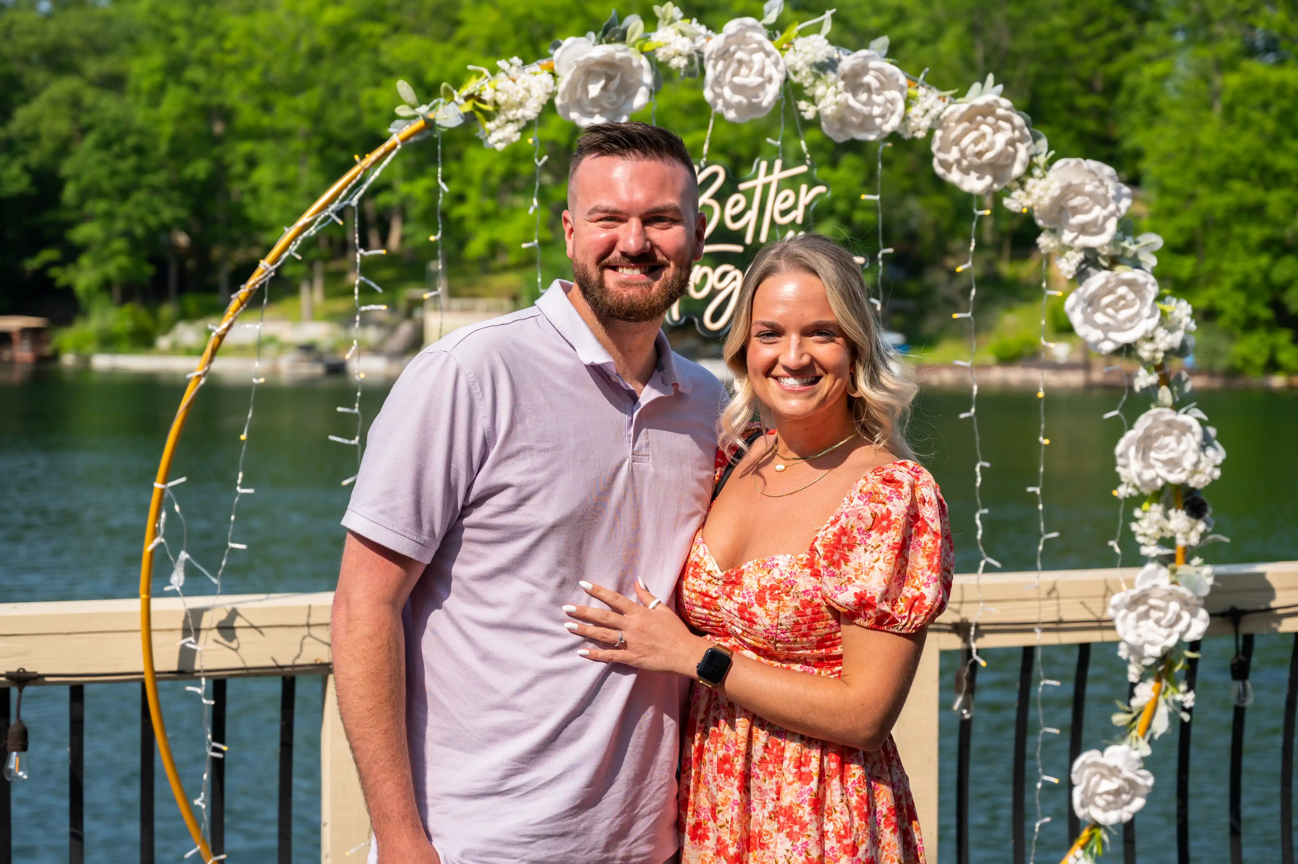 A smiling couple embracing in front of a floral arch with the words "better together" on a lakeside deck, indicative of a celebration or special event.