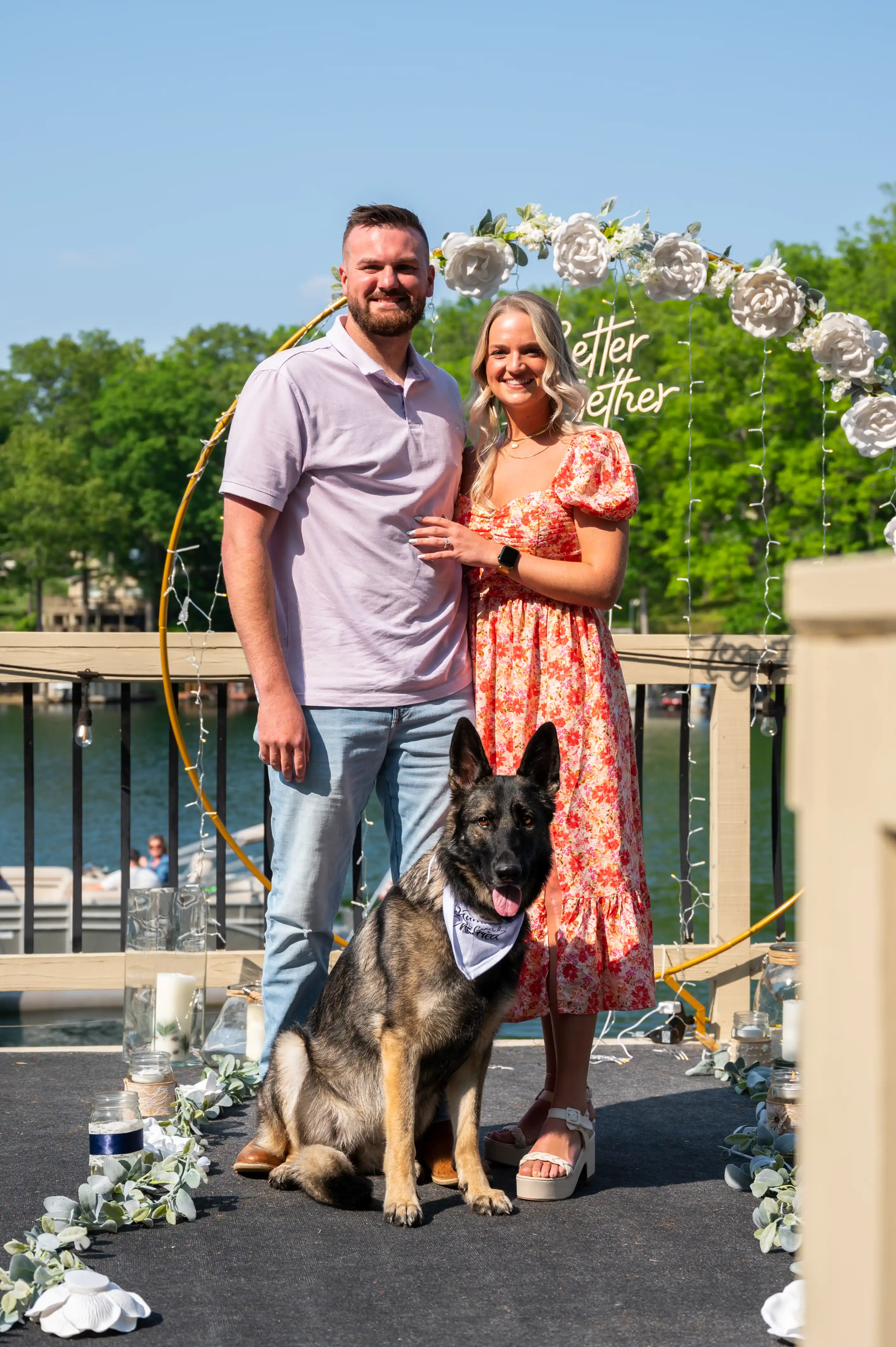 A smiling couple posing with a German Shepherd dog on a bridge with decorative foliage and flowers in the background.