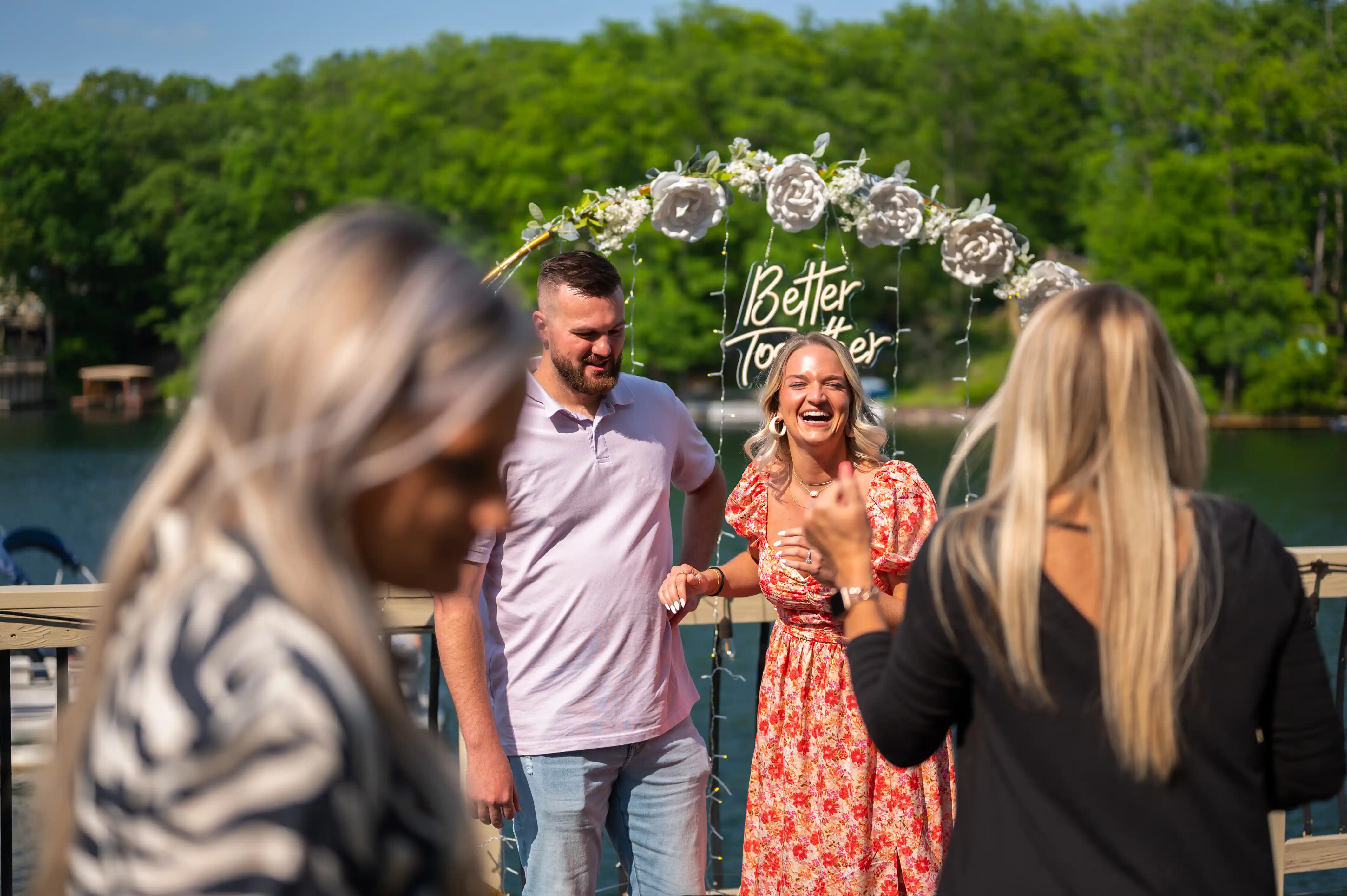 A couple is engaging with a woman applauding them in the foreground, with an arch decorated with flowers and "Better Together" sign in the background by a lake.