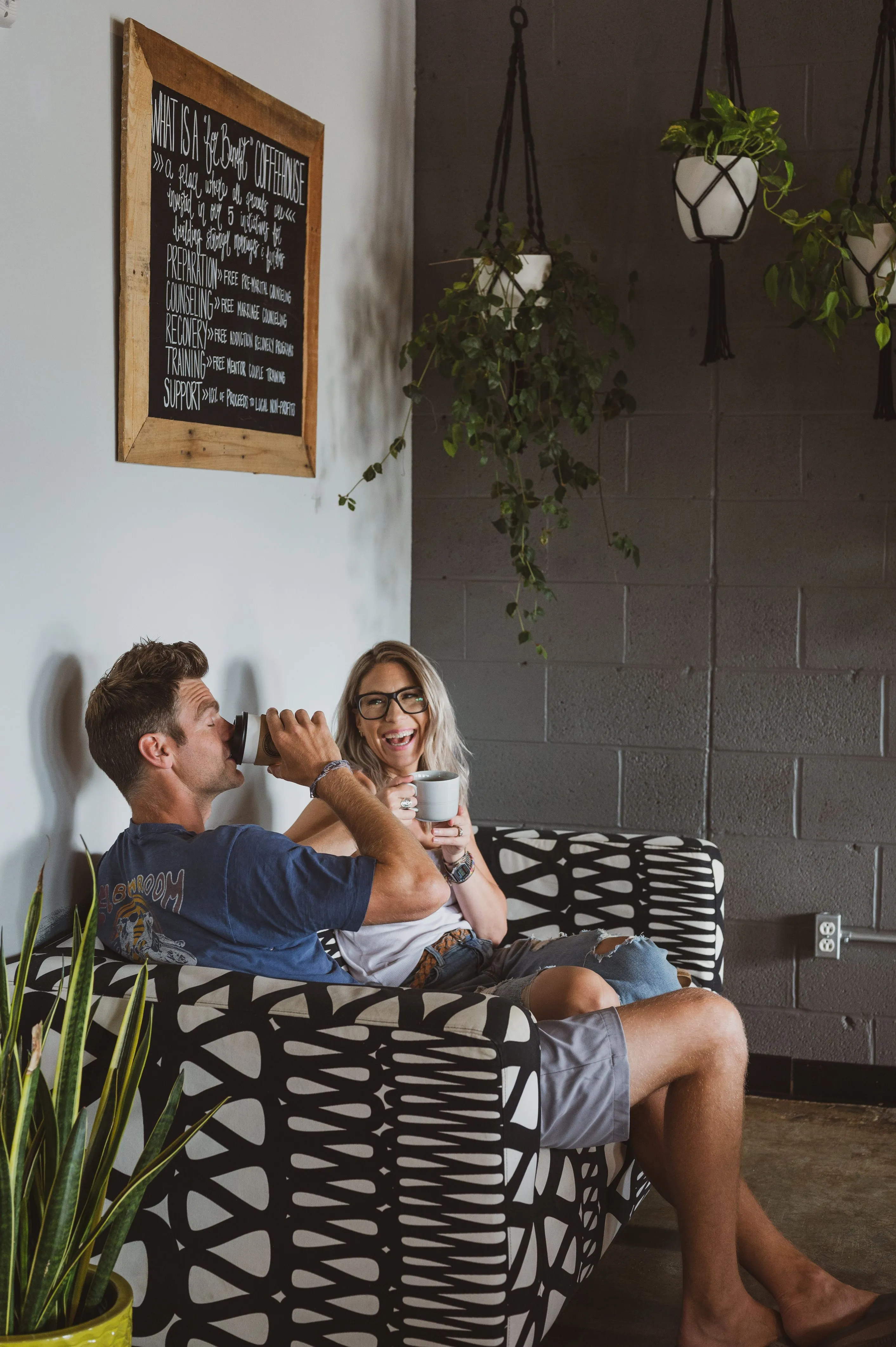 A man and a woman sitting on a patterned couch, enjoying coffee together in a cozy coffee shop setting with hanging plants and a chalkboard menu in the background.