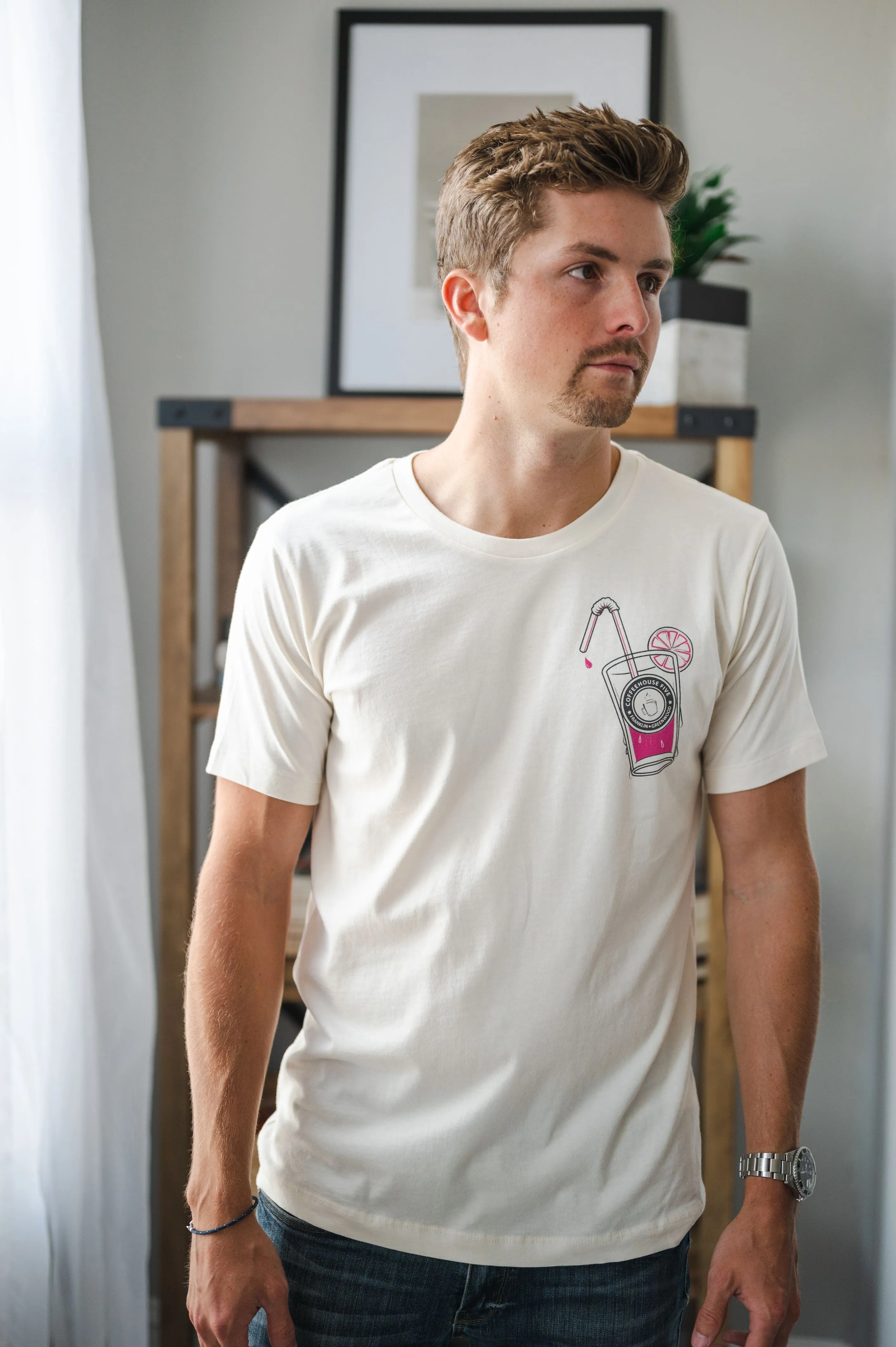 Man in a casual pose wearing a white t-shirt with a pink camera graphic design, standing indoors near a window with soft light.
