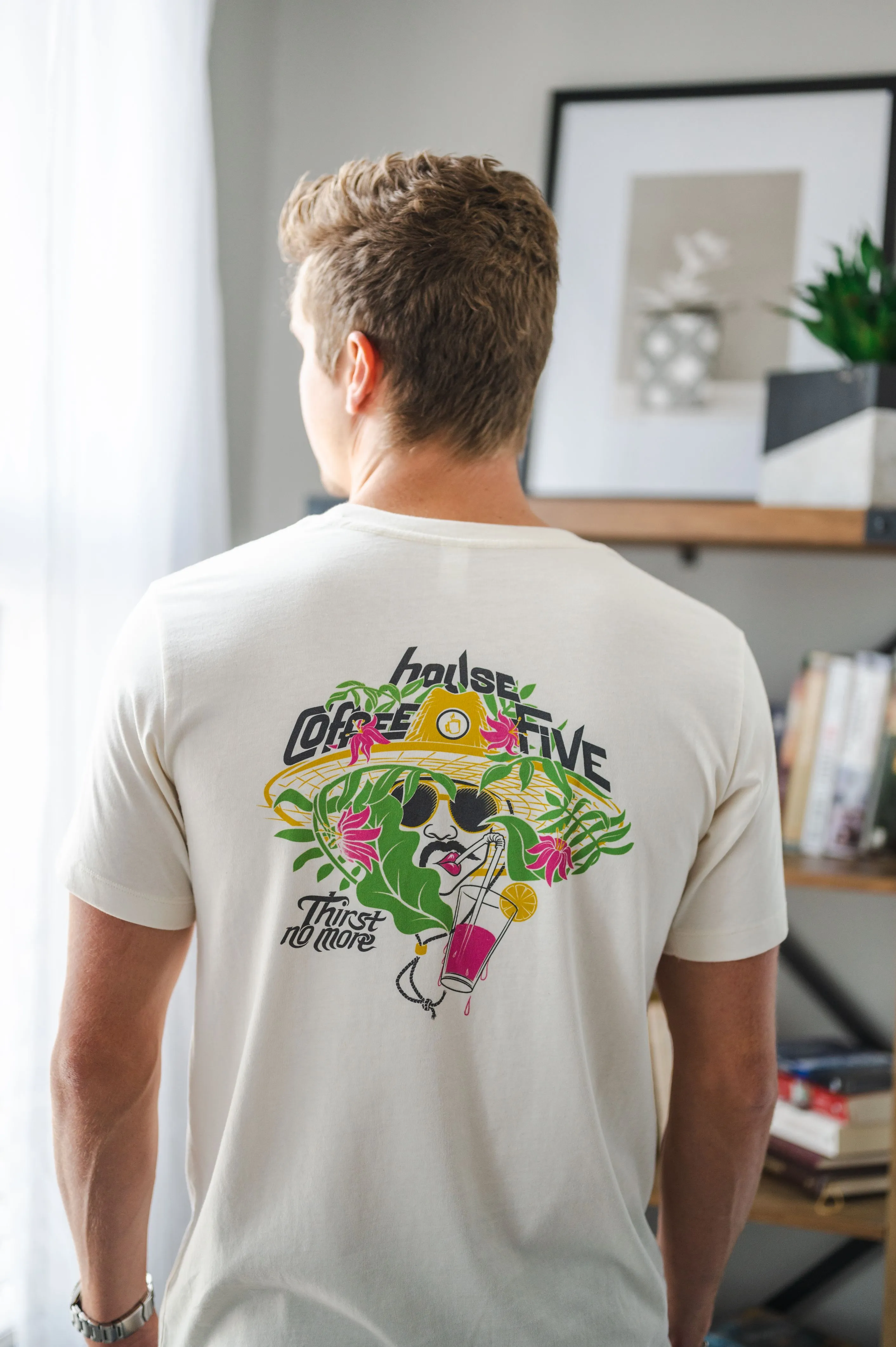 Rear view of a person wearing a white t-shirt with a colorful graphic print, standing in a room with a bookshelf and framed art in the background.
