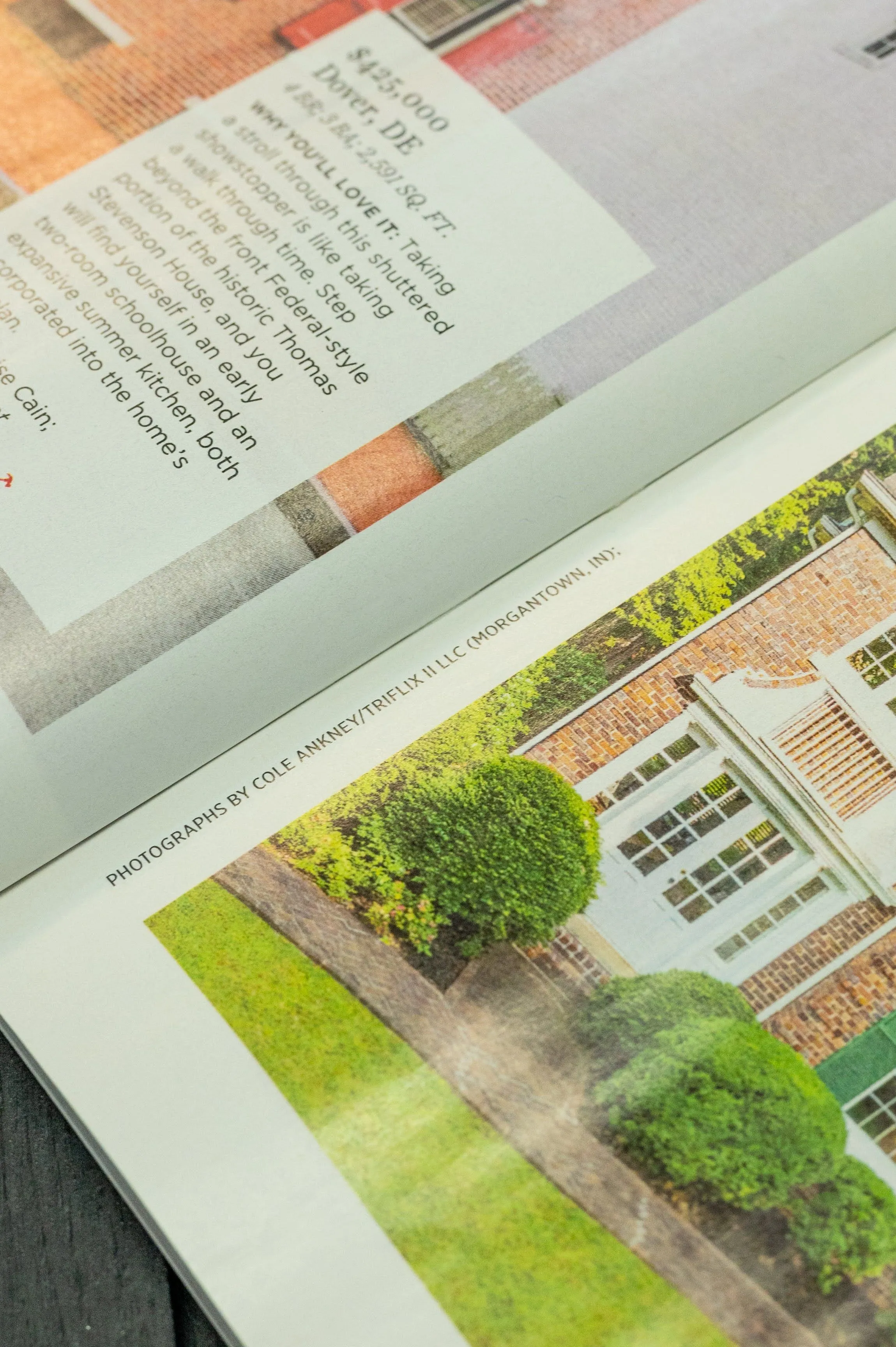 Close-up view of an open magazine featuring a real estate listing with a photograph of a brick house and green shrubbery, and a credit line for the photographer at the bottom of the page.