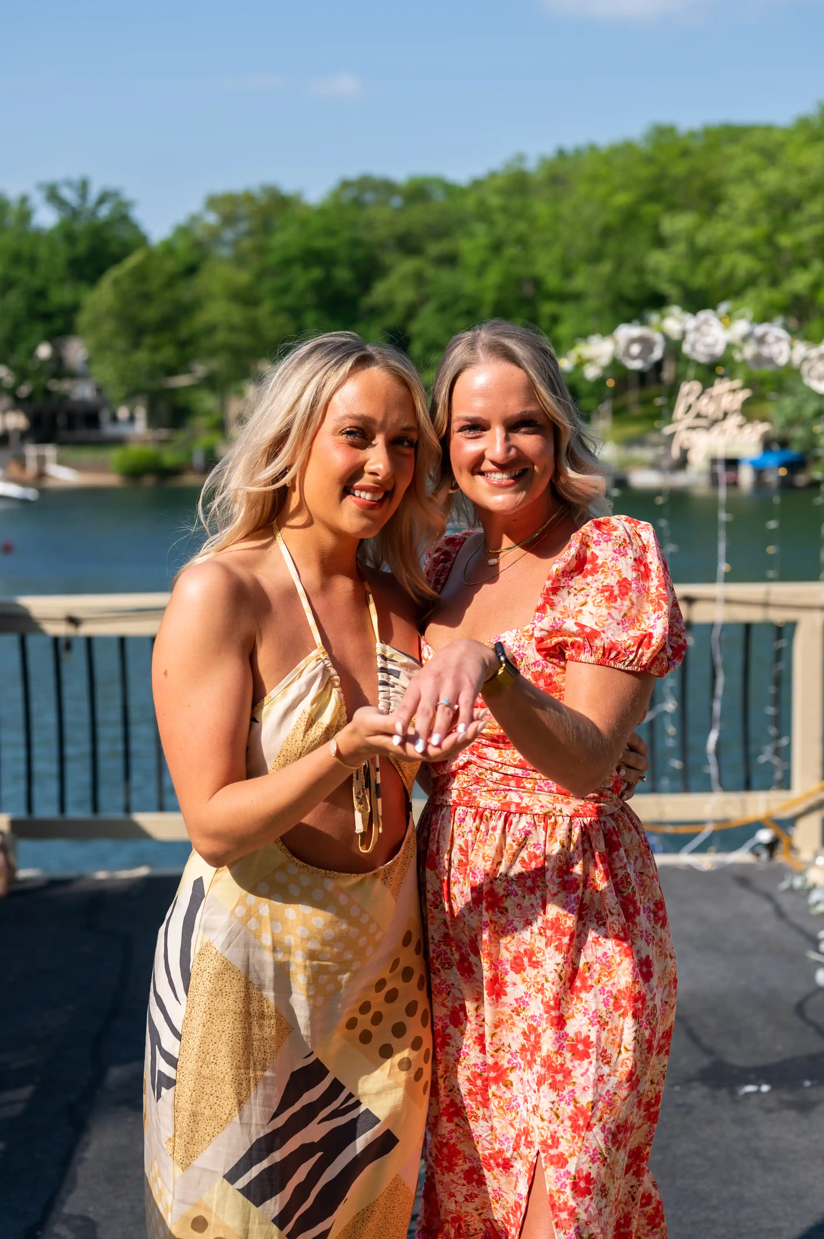 Two smiling women in summer dresses standing on a deck with a lake and trees in the background.