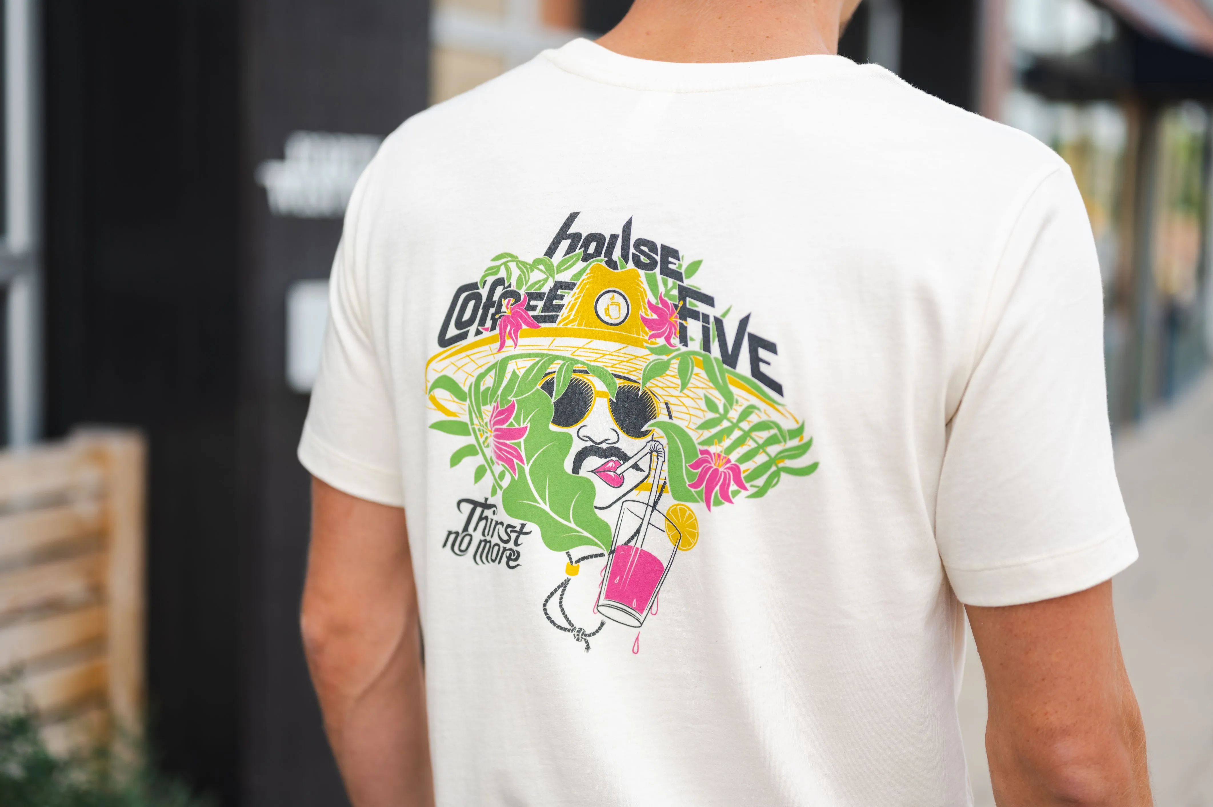 Person wearing a white t-shirt with a colorful graphic of a character in a sombrero surrounded by tropical flora and holding a drink with the text "house coffee five - Thirst no more".